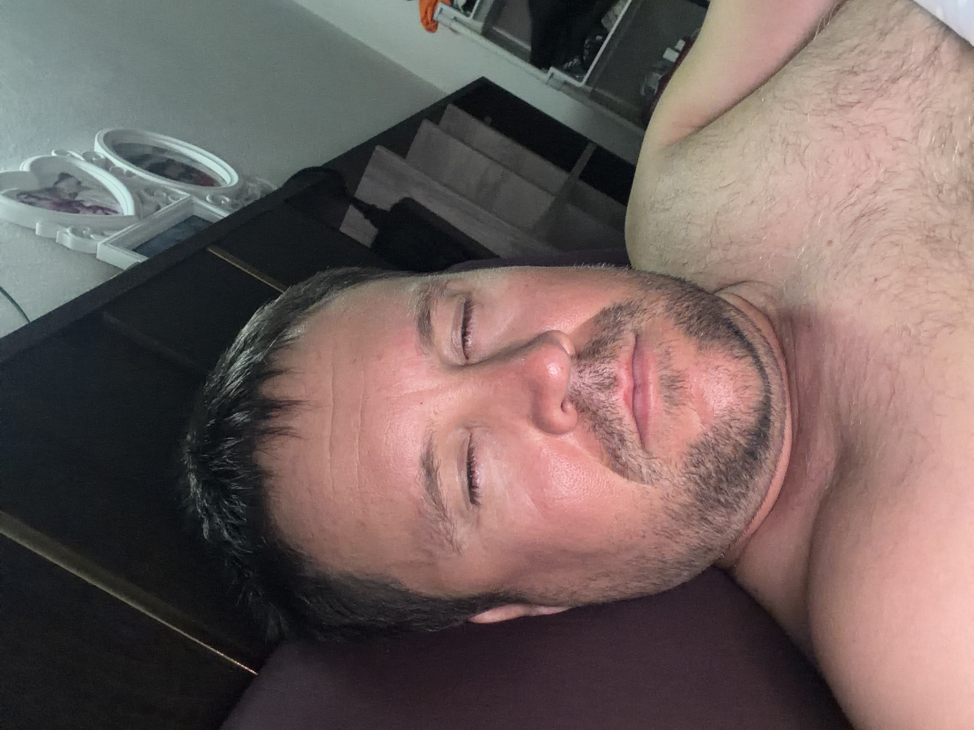 The image is a close-up, overhead selfie of a man lying down. He is shirtless and has his eyes closed. The man has short, dark hair and stubble on his face. The background is dark, with a hint of a shelf unit and some framed pictures on a wall. The overall lighting suggests an indoor setting, possibly a bedroom. There are no visible clues or features within the image that would indicate the specific location of Baan Mae Nam, Thailand.