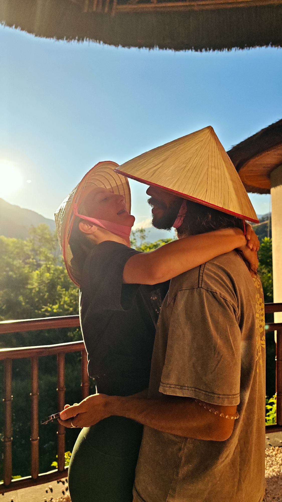 This outdoor portrait captures a joyful and intimate moment between a couple embracing on what appears to be a balcony or terrace in Mu Cang Chai, Vietnam. Both individuals are wearing traditional Vietnamese conical hats (nón lá); the man's hat has a distinct red rim, and the woman's is secured with a pink chin strap.
The woman, positioned on the left, is dressed in a black top and dark green leggings. She looks upwards at the man with a wide, happy expression, suggesting laughter or a broad smile. Her left hand holds a small, dark object, possibly a mobile phone or vape pen. The man, on the right, has a beard and long hair, wearing a light brown or grey t-shirt with a faint pattern on the back. He has a beaded bracelet on his right arm and a ring on his left ring finger. His left arm is wrapped affectionately around the woman's back.
The scene is bathed in warm, golden sunlight from the left, indicative of the golden hour, either late afternoon or early morning. This strong backlighting highlights the subjects and creates a vibrant atmosphere against a clear, bright blue sky. In the background, lush green mountains and trees provide a natural, scenic backdrop typical of Mu Cang Chai. A sturdy wooden railing runs across the foreground and mid-ground. Above and to the right, a structure with a thatched roof is partially visible, suggesting a traditional building or resort. The image conveys strong emotions of affection, happiness, and connection between the couple. No discernible text is visible within the frame.
