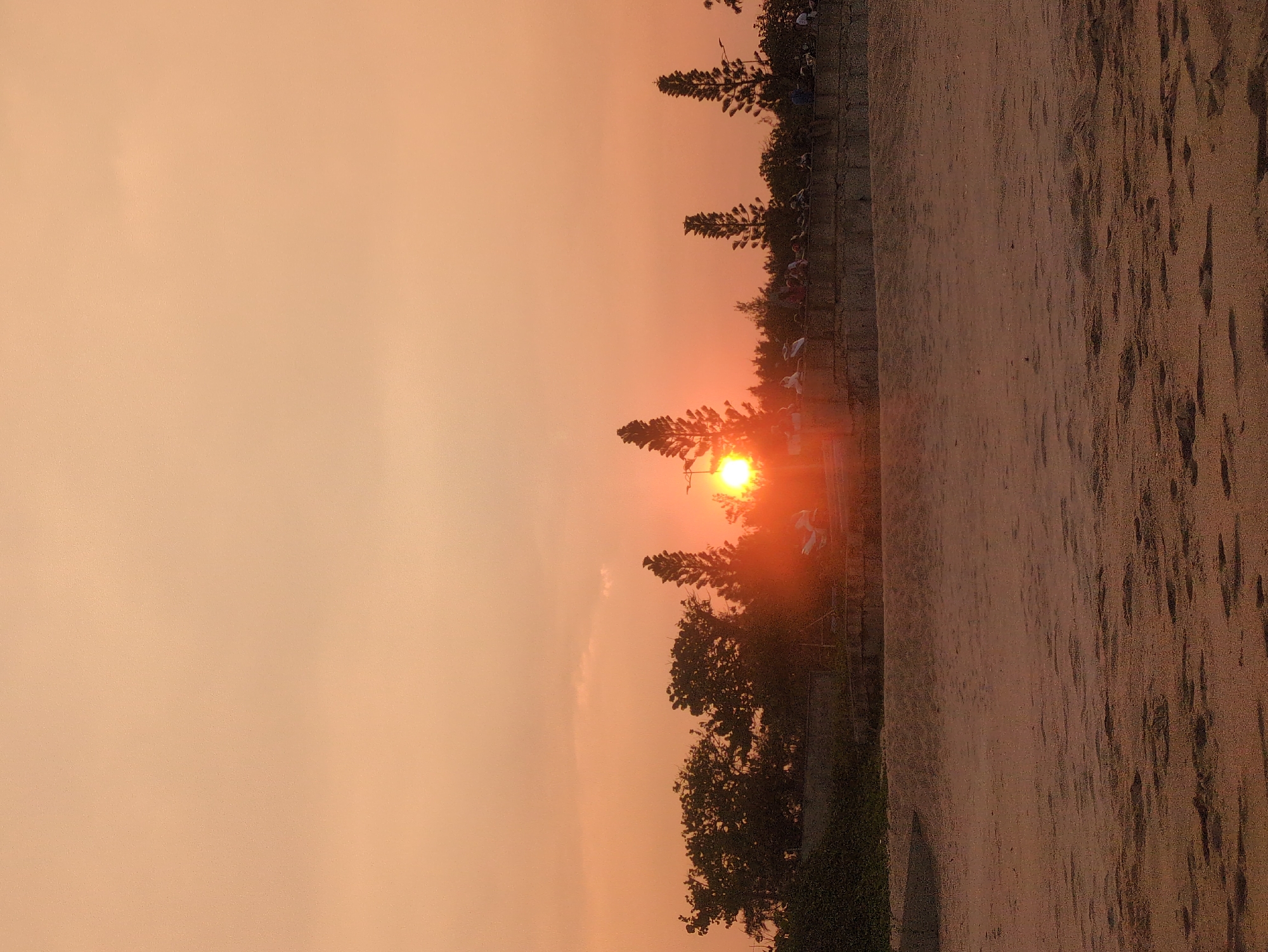 At sunset in Tuy Hòa, Vietnam, the orange sun is partially obscured by trees along the edge of a beach. A crowd of people is visible, partially obscured by trees. The beach is sandy.