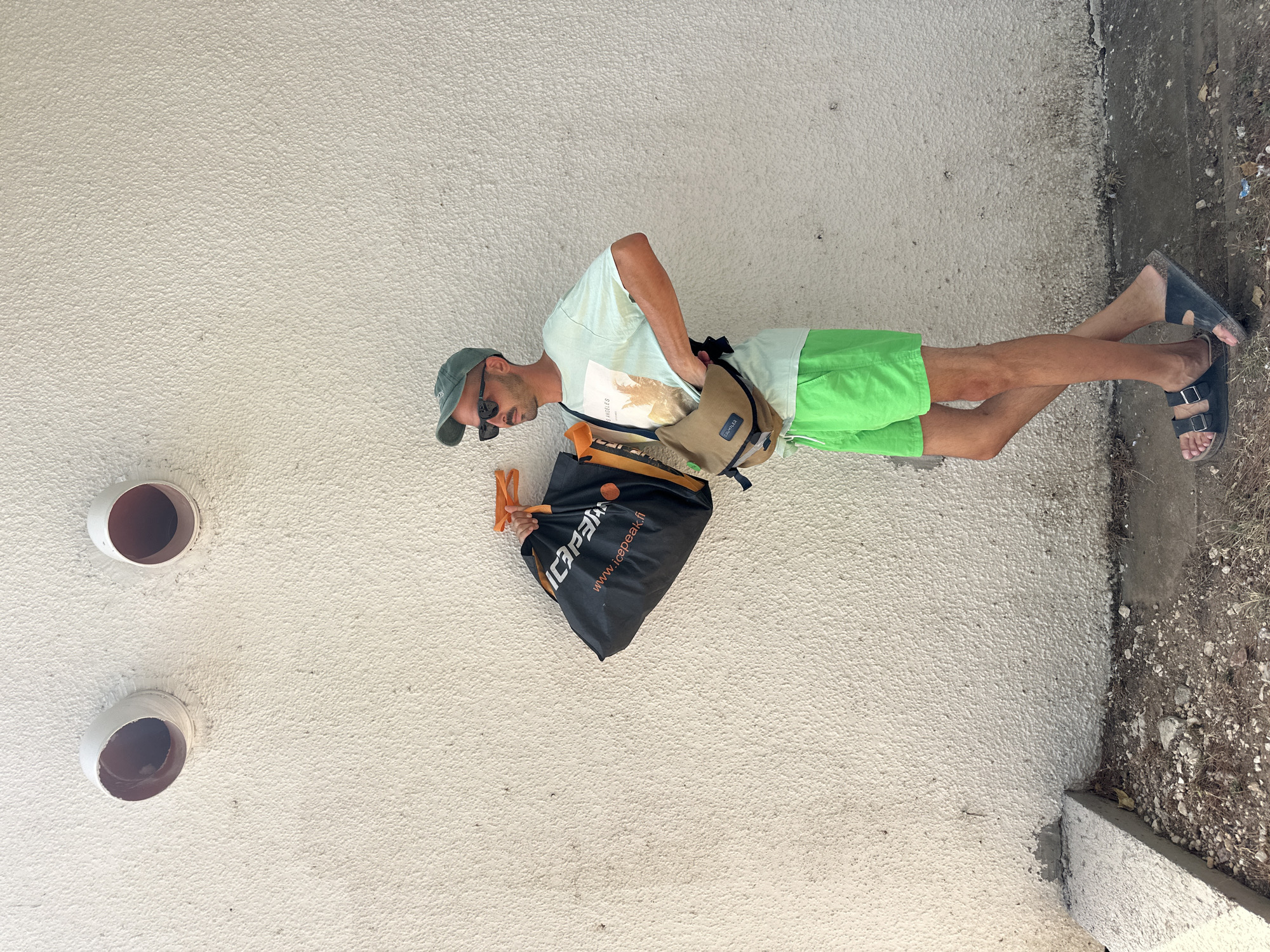 A man stands against a textured white wall, holding a black bag with orange handles and a tan messenger bag slung across his chest. He wears a light green t-shirt with a graphic print, bright green shorts, a green baseball cap, and black sandals. The wall has two circular openings near the top. The ground in front of the wall is a mix of concrete and gravel. The scene appears to be an outdoor urban setting.