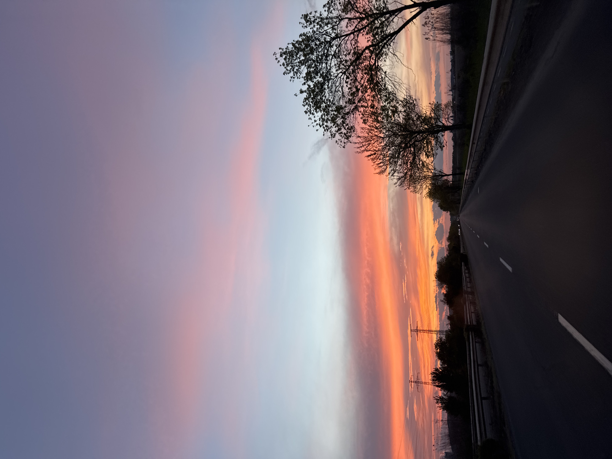 The image depicts a twilight scene in Burgas, Bulgaria, featuring a road running into the distance. The sky exhibits vibrant hues of pink, orange, and blue, likely during sunset or sunrise. A few silhouetted trees are visible along the roadside. Power lines are located next to the road. The road surface is dark and asphalt, with white lane markings.