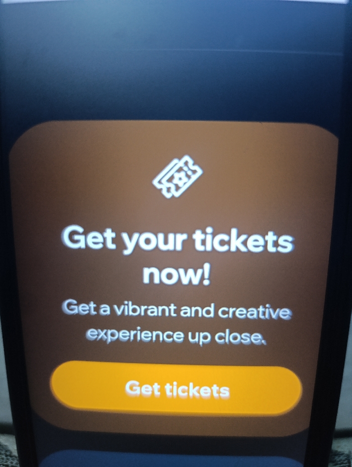 The image shows a smartphone displaying a web page. The webpage is a call to action for a user to buy tickets. It is a brown background with the words "Get your tickets now!" in white text, with a white icon of a ticket above. The next line of text reads, "Get a vibrant and creative experience up close." There is a large, yellow button at the bottom of the screen that reads "Get tickets" in white. There are no people or objects present in the scene other than the smartphone. The location of the smartphone is Kecamatan Arjasa, Indonesia, based on the provided coordinates. The smartphone is likely being held by someone, but this is not clear from the image. The image provides no details about the specific event for which tickets can be purchased.