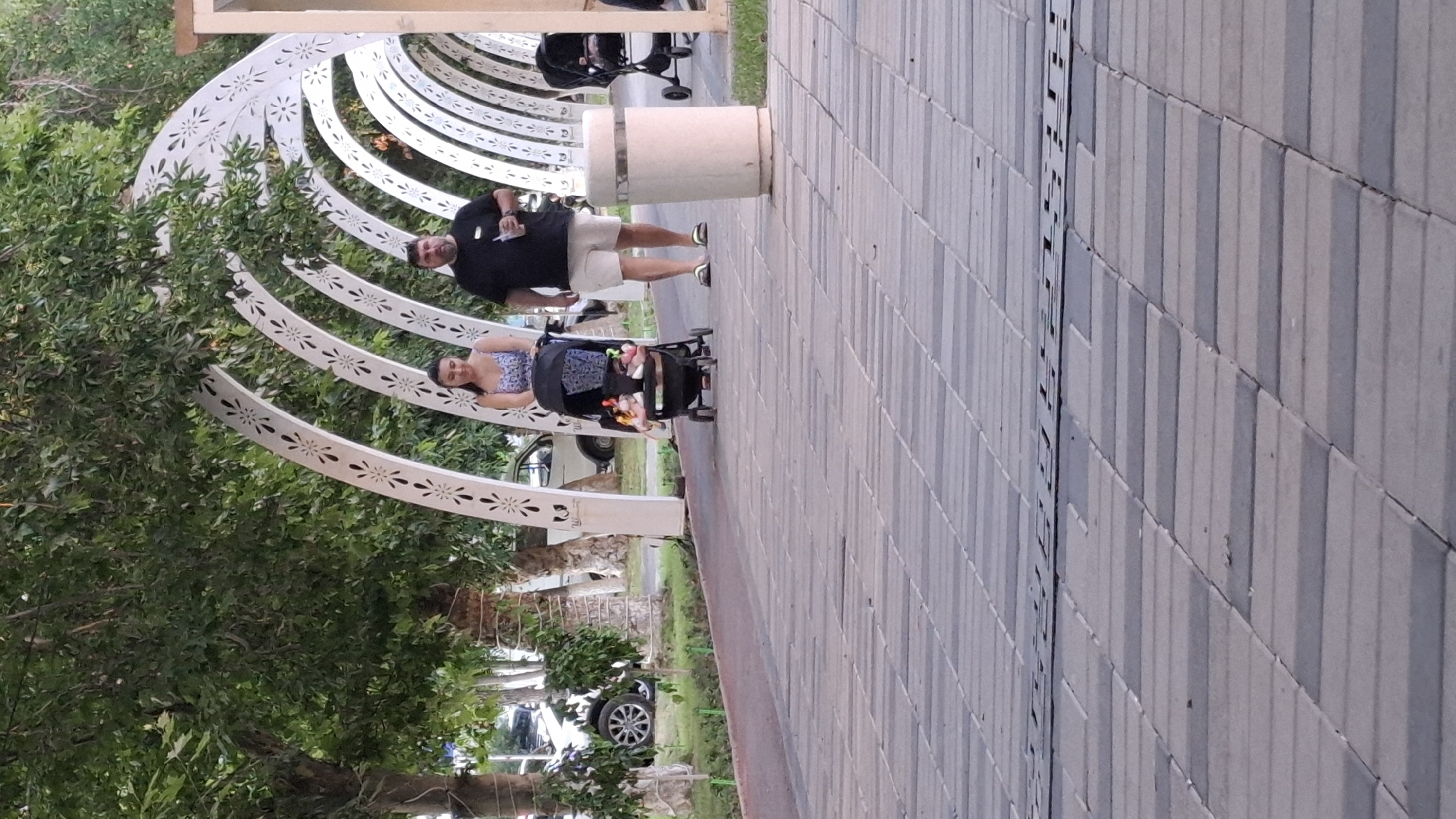 The image depicts a walkway in Shymkent, Kazakhstan, featuring a decorative arched structure covered in foliage. A man and a woman are walking along the path, with the woman pushing a stroller. The man is holding a drink in his hand. In the background, there are trees and cars. The walkway is paved with rectangular stones. The overall scene appears to be a public space, possibly a park or promenade, on a sunny day.