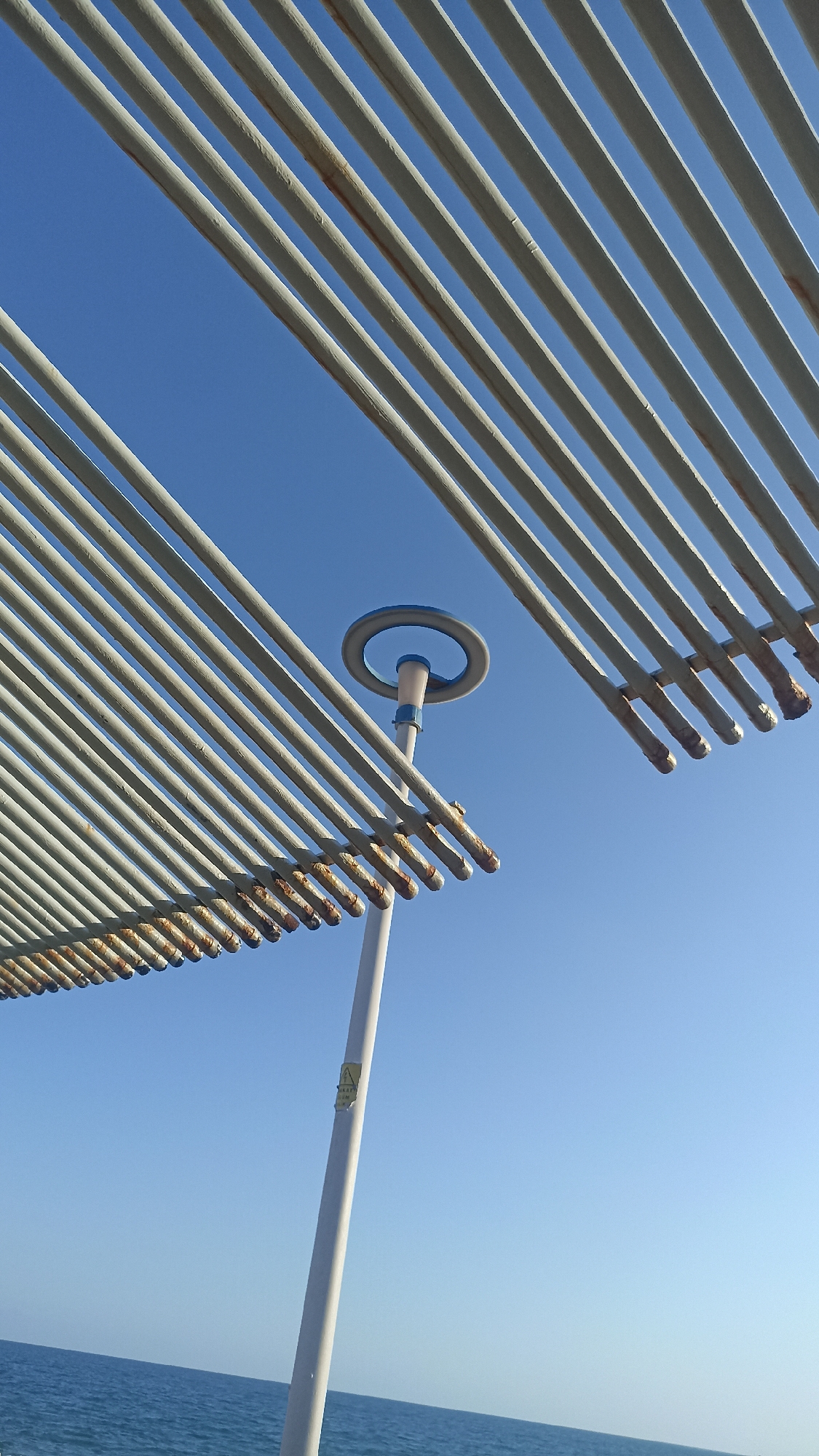 This is a low-angle, outdoor, vertical, eye-level shot of a modern outdoor structure with a clear blue sky and a body of water in the background. The structure appears to be part of a beachside or seaside amenity, possibly a covered walkway or seating area, given the presence of the ocean.
The primary subjects are the parallel white metal beams of the structure, which create a striped pattern against the sky. These beams are angled from the bottom left and top right of the frame, converging towards the center. Some of the beams show signs of rust on their ends, suggesting exposure to the elements.
In the upper central part of the image, a white pole rises, supporting a circular element with a blue outline. This could be a decorative feature, part of a lighting fixture, or a signage holder. A yellow triangular sticker with black text is visible on the pole, but the text is too small to be legible.
The setting is clearly outdoors, with a vast expanse of clear blue sky occupying most of the frame. Below the structure, a calm, deep blue ocean stretches across the bottom of the image. The lighting suggests it is daytime, likely midday or afternoon, with bright sunshine. The weather appears to be clear and sunny.
There are no people visible in the image, and no specific activity is taking place. The overall mood is one of quiet and perhaps a slight sense of neglect due to the visible rust, contrasting with the otherwise clean and modern design of the structure and the serene natural backdrop. The location cues, such as the ocean and the architectural style, are consistent with a seaside resort or public area.