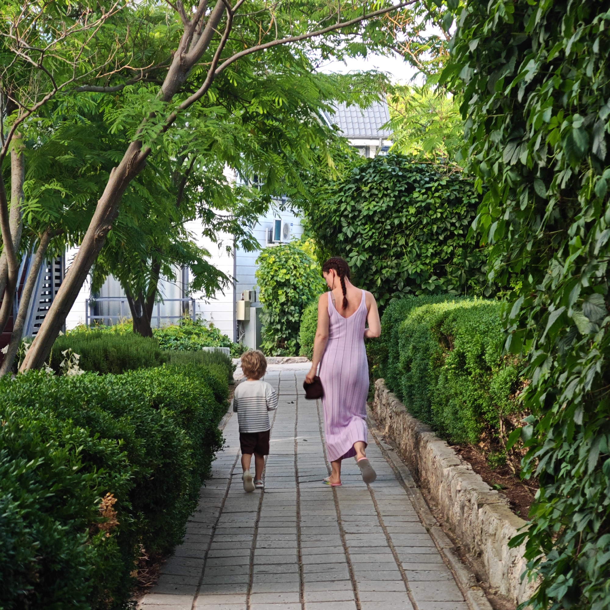 In the city of Мысовское сельское поселение, Ukraine, a woman and a child walk down a paved pathway lined with lush green bushes and trees. The woman, wearing a light purple dress, walks ahead of the child, who is dressed in a striped shirt and brown shorts. The pathway is made of rectangular stone tiles. On the left side of the path, there are manicured bushes and trees with visible trunks. On the right, a dense green vine covers a stone wall. In the background, a white building with windows is partially visible through the foliage, and a portion of a grey tiled roof can be seen. The scene appears to be a park or a garden area within the settlement.