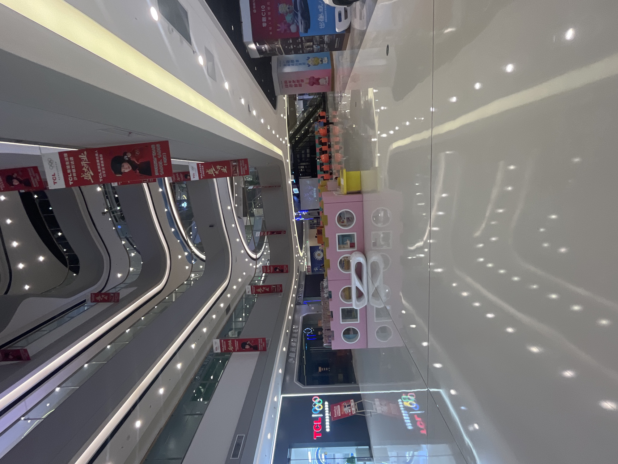The image shows the interior of a multi-story shopping mall, likely located in Shapingba District, China, given the context provided. The mall features a modern architectural design with curved balconies, bright lighting from numerous ceiling fixtures, and a reflective, glossy floor that creates numerous light reflections. Red banners with Chinese text and images are hung vertically from different levels of the mall. One banner prominently features the TCL logo and Olympic rings, along with images of a person and promotional text. A TCL store is visible on the left side of the frame with its logo and display. In the mid-ground, there is a children's play area with pink, block-like structures containing circular openings, and a white modern sculpture. Further back, escalators are visible, and various displays and store fronts line the walkways. Several people are present, including a mall employee in uniform standing near the escalators, and other individuals in casual attire walking through the mall. The overall impression is of a clean, well-lit, and active commercial space.