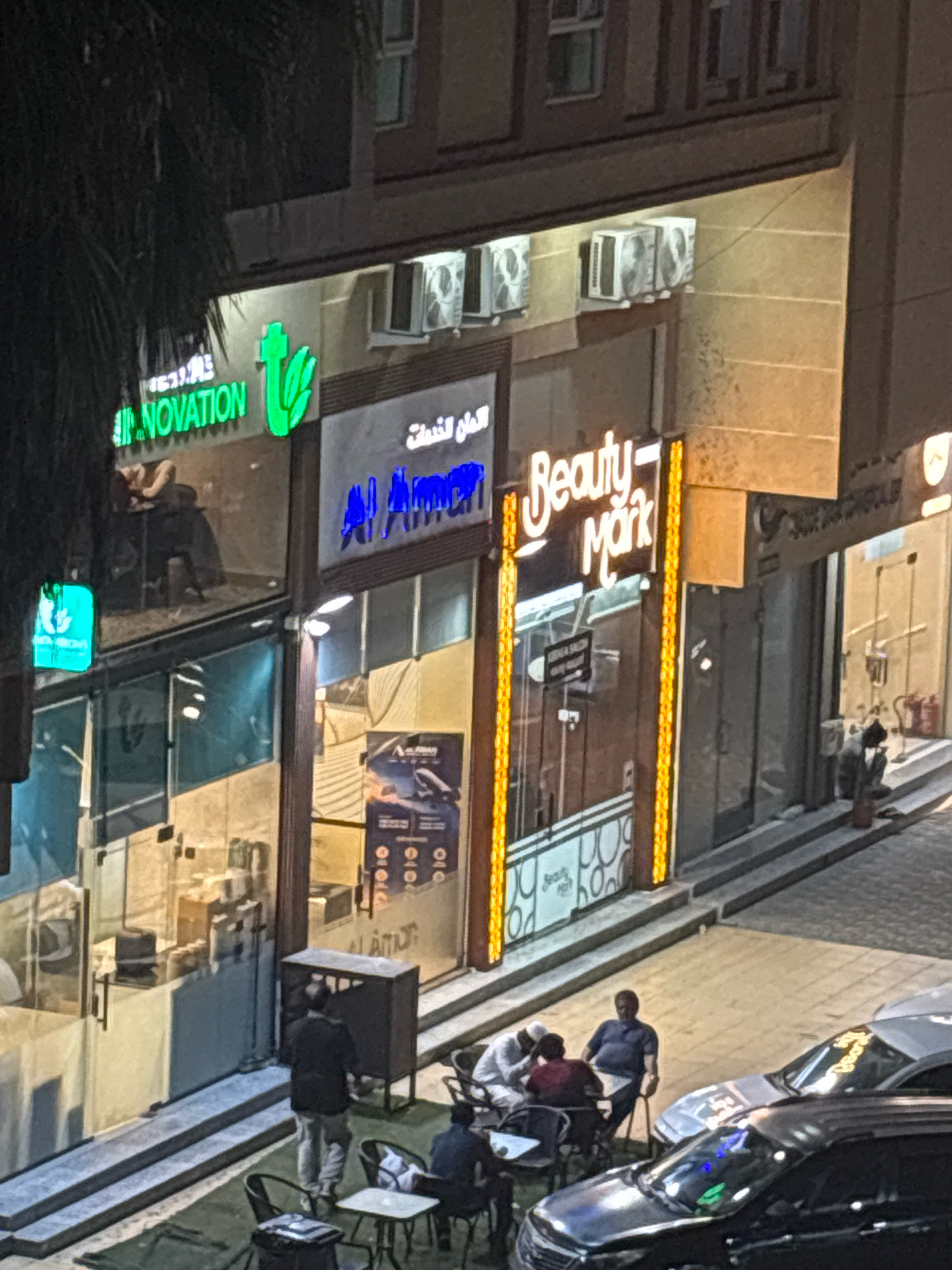 The image depicts a street-level view of businesses in Dammam, Saudi Arabia, at dusk or night. The storefronts are illuminated, highlighting the signage. One prominent sign displays "Beauty Mark" in white illuminated script with decorative yellow lighting borders. To the left, another business has a green illuminated sign with the text "INNOVATION" and a stylized green leaf symbol. Next to it, a sign in Arabic script is visible in blue neon. The buildings have multiple stories with visible air conditioning units mounted on the exterior walls.
In front of the businesses, there is a seating area with several tables and chairs set up on a green surface, resembling artificial grass. People are seated at these tables. One man in a dark shirt is seated at a table. Two other individuals, one wearing white head covering, are seated facing each other. Another man in a blue polo shirt is seated at a separate table.
A dark SUV is parked in the foreground, with another silver vehicle visible behind it. The street and sidewalk area are paved. Shadows are cast from the buildings and lighting, indicating the time of day. A person in a black jacket and light-colored pants is walking away from the camera, near a dark gray cabinet-like structure. Several storefront windows reflect the surrounding lights and environment.