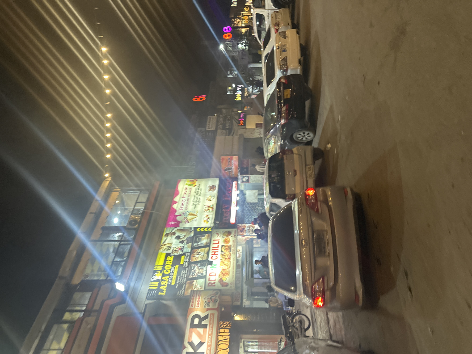 The image depicts a street scene at night in Quetta City Tehsil, Pakistan. Several cars are parked along the right side of the street. On the left side, a building is lined with numerous brightly lit commercial signs for businesses including "LASA CORE," "RED'N CHILLI," "Frosty Freeze," "KR COME," and "BeeBerry." People are visible in front of the businesses. Overhead, strings of lights stretch across the street.