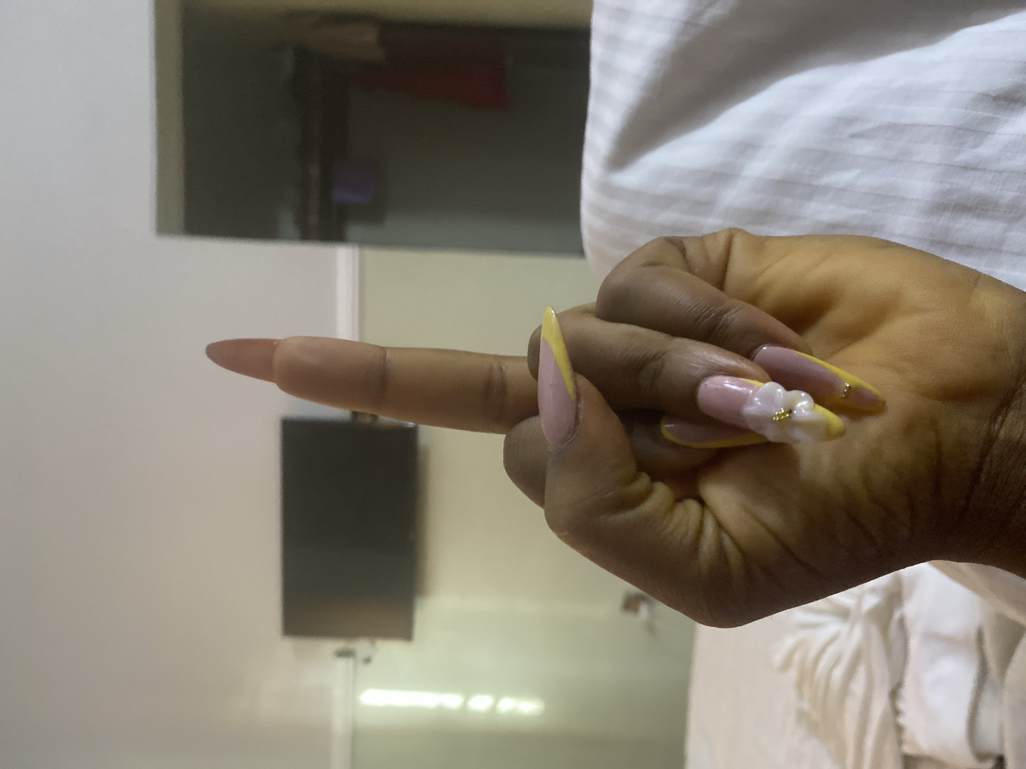 In Howayi, Nigeria, a close-up shot shows a person's left hand with elaborate, long, almond-shaped fingernails adorned with pink, yellow, and white floral designs. The hand is positioned in the foreground, with the index finger extended upwards. In the background, a television is mounted on a wall, and a closet door is visible in an adjacent room. The setting appears to be a bedroom, with a white, textured bedsheet dominating the lower portion of the frame. The lighting suggests an indoor environment, possibly a hotel room.