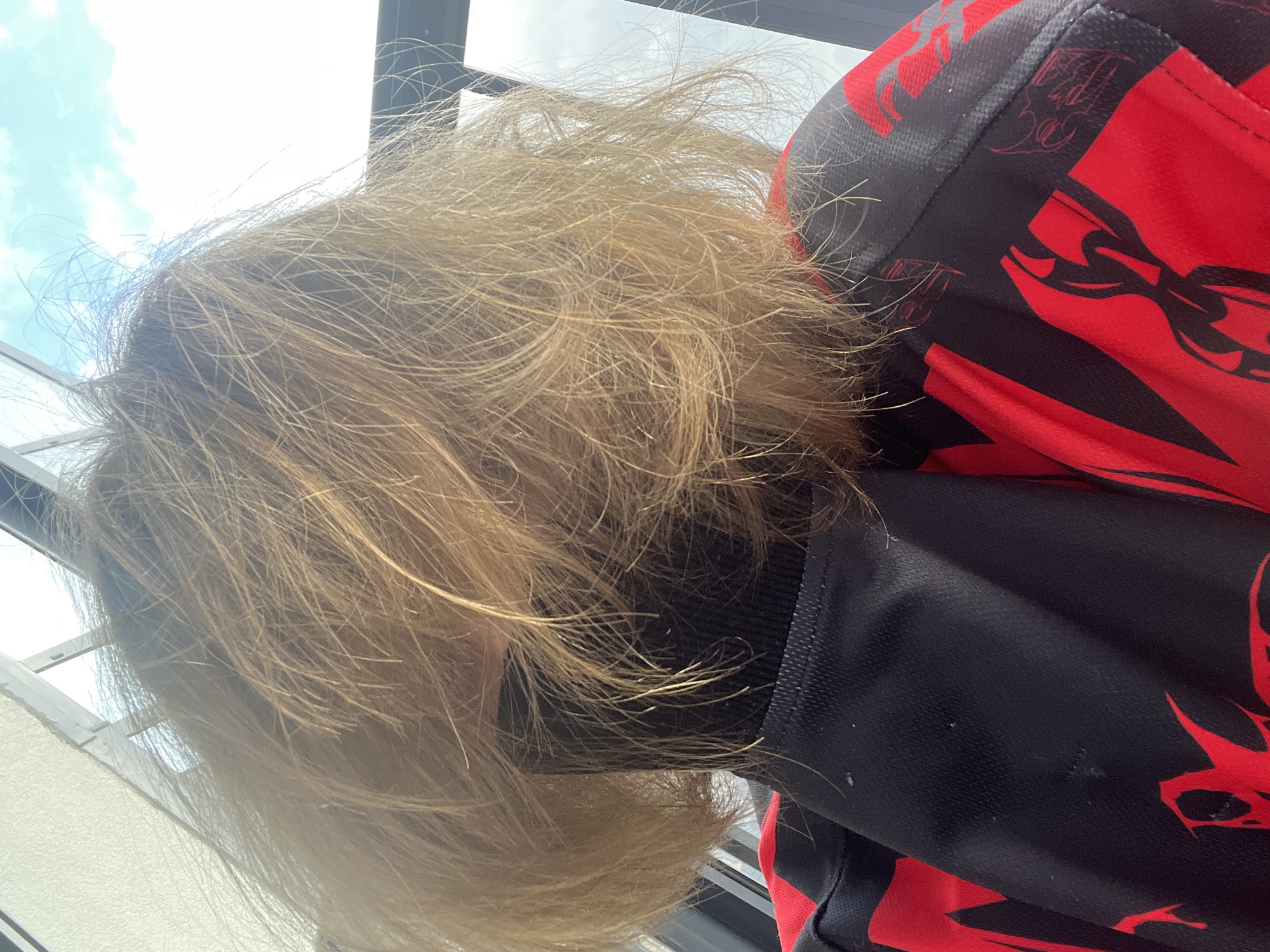 A close-up, rotated image captures the head and shoulder of a person with light blonde hair. The hair appears somewhat disheveled or windswept, with many individual strands visible against the bright background. The person is wearing a long-sleeved shirt that is primarily black with striking, stylized red patterns, possibly abstract shapes or chains. The fabric of the shirt appears to be a performance material.
The background is brightly lit, showing a clear, slightly cloudy sky, indicating daytime. Architectural elements, possibly window frames or parts of a building, are faintly visible against the sky. The perspective is from an overhead or slightly upward angle, looking down or across the person's head, which is largely obscured by their hair. No specific activity is discernible, and no text is visible on the clothing or in the background. The scene is well-lit, highlighting the hair. The location context is Saint Petersburg, Russia.