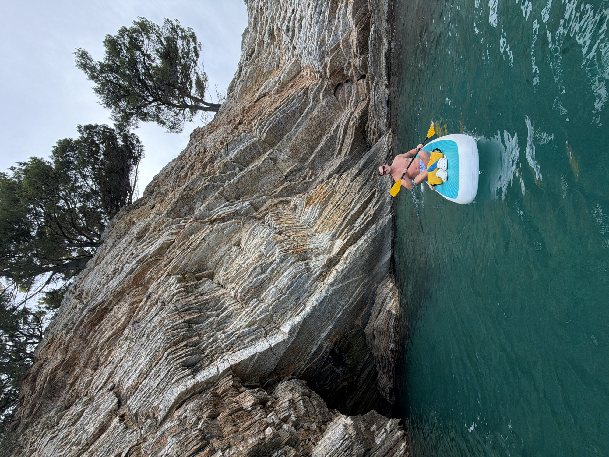 A shirtless man wearing blue and white patterned swim trunks and sunglasses is paddleboarding in the turquoise waters of Petrovac centar, Montenegro. He is holding a yellow paddle. The paddleboard is white with a light blue accent on the top and has two yellow flotation devices strapped to it. The man is positioned on the right side of the frame, while a steep, rocky cliff face dominates the left side. Trees are growing out of the top of the cliff. The sky is overcast. The overall impression is one of a leisurely day spent enjoying the natural beauty of the Montenegrin coast.