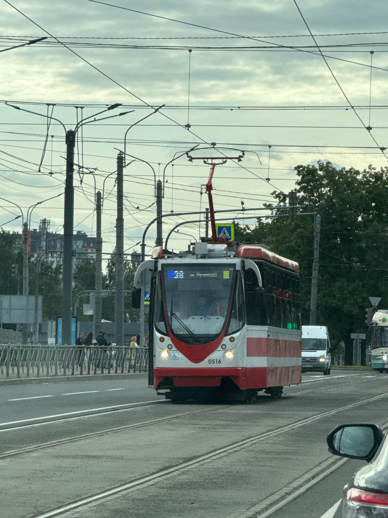 A red and white tram, numbered 0516, is traveling on tram tracks on a cloudy day. The tram has a pantograph connected to overhead wires. In the background, there are several tall buildings, trees, a white van, and another tram. A reflective car mirror is visible in the lower right corner of the image. The scene appears to be a street in Saint Petersburg, Russia, given the architecture and the presence of a tram.