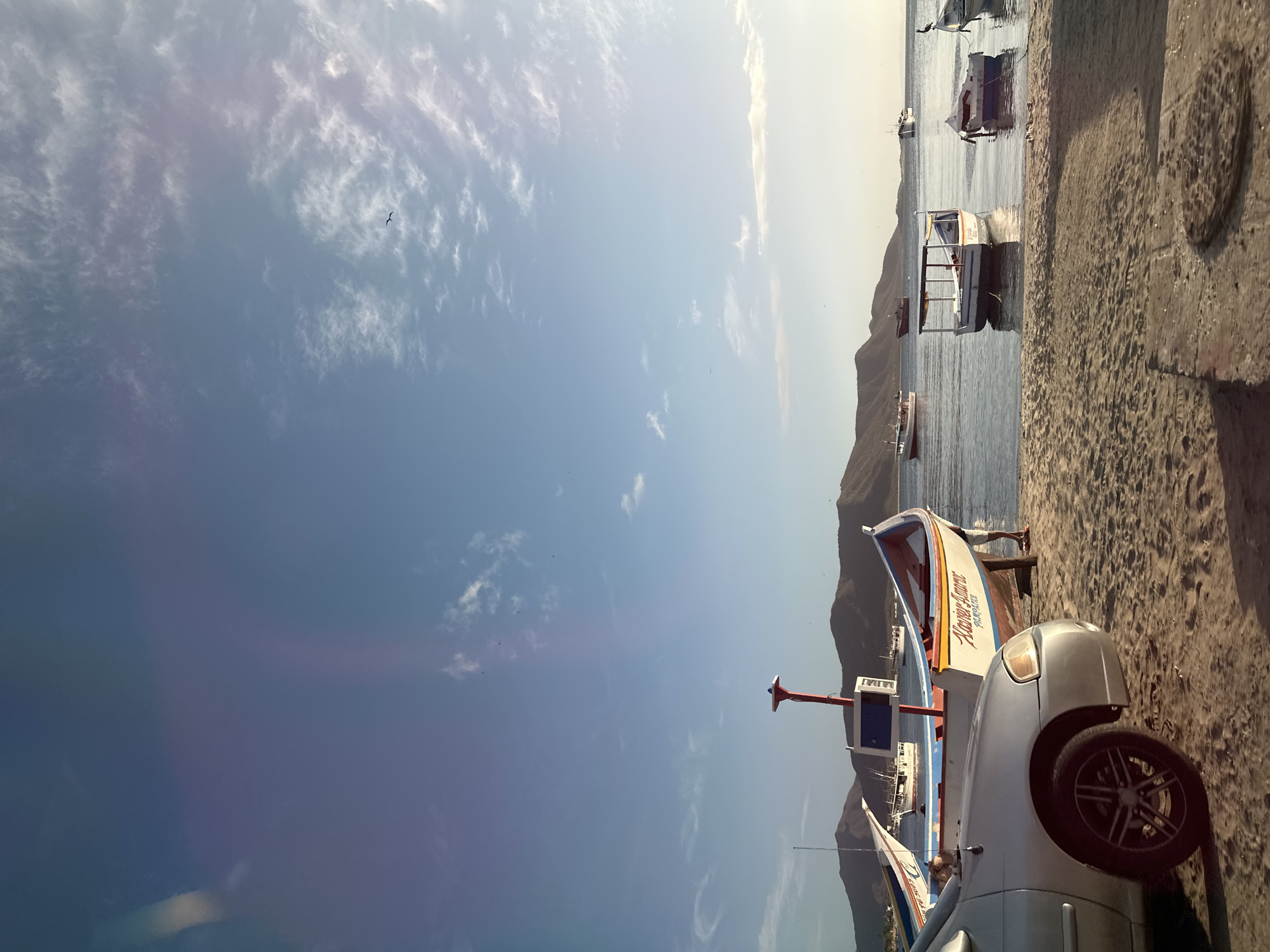 A photograph taken from a slightly low angle on a beach in Juan Griego, Venezuela, captures a serene coastal scene under a clear blue sky with sparse white clouds. In the foreground, a silver car is parked on the sandy shore, its front left wheel clearly visible. Adjacent to the car, a colorful fishing boat with a red and white hull and a blue trim rests on a wooden stand on the sand. The name "XAVIER AMOR" is painted on its side. Further out in the calm, clear water, several other fishing boats are moored or gently floating. In the background, a range of rolling, verdant mountains forms the horizon. The sunlight casts shadows on the sand and highlights the water's surface, suggesting it is late afternoon or early morning.