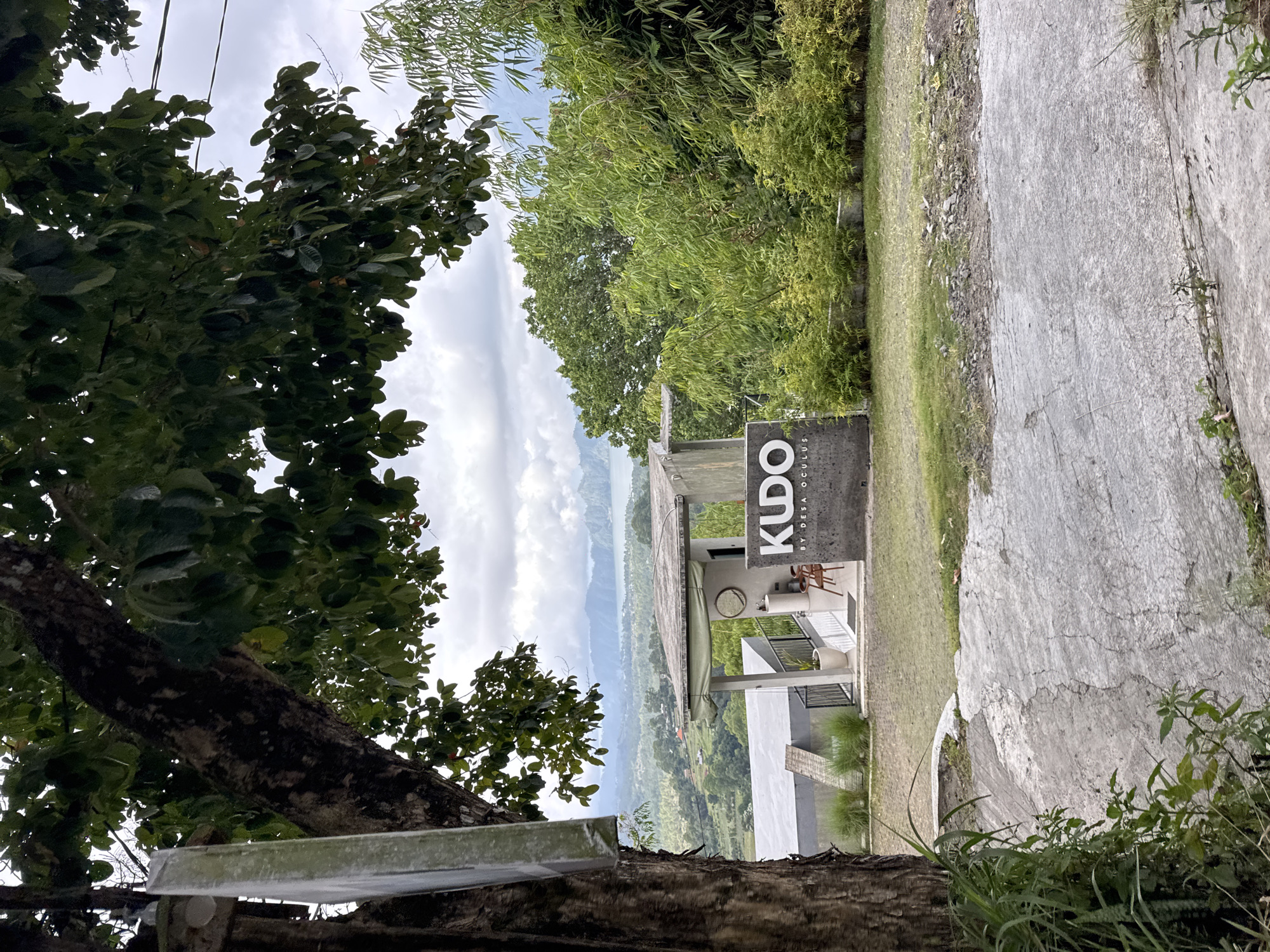 The image shows a landscape view in Kintamani, Bali, Indonesia. In the foreground, a large tree trunk with dark bark and green leaves partially obscures the view on the left. A rough, cracked concrete path leads towards the middle ground.
Dominating the mid-ground is a low, modern building with a gray facade, featuring an open-air patio area. The building has a flat roof and large windows. A dark gray sign with white lettering that reads "KUDO BY DESA OCULUS" is positioned in front of the building. Two wooden chairs and a small round table are visible on the patio, along with a potted plant.
Behind the building, lush green foliage, including bamboo plants and other trees, creates a dense natural backdrop. In the distance, a large body of water, likely Lake Batur, is visible, surrounded by rolling hills and mountains under a cloudy sky. The overall scene suggests a scenic overlook or a cafe with a view of the natural landscape. There are no people visible in the image.