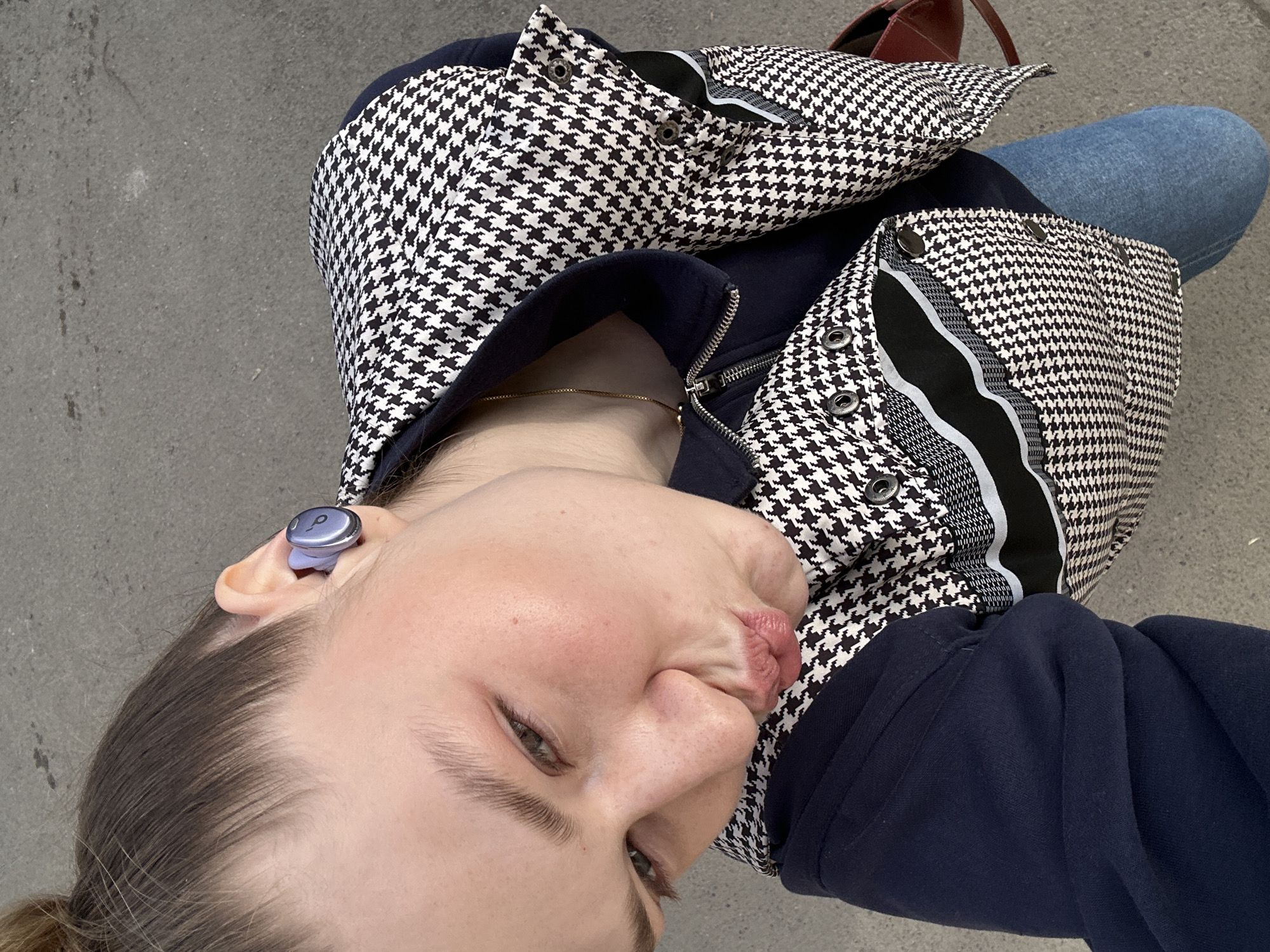 A person is sitting outside in an unknown location. The person is wearing a dark blue zip-up jacket with a houndstooth patterned fabric on the upper part and along the trim. A light purple earbud is visible in their left ear. The person is also wearing blue jeans. Part of a red or maroon bag is visible in the upper right corner of the image. The background appears to be concrete. The photo was not taken in Kazan, Russia.