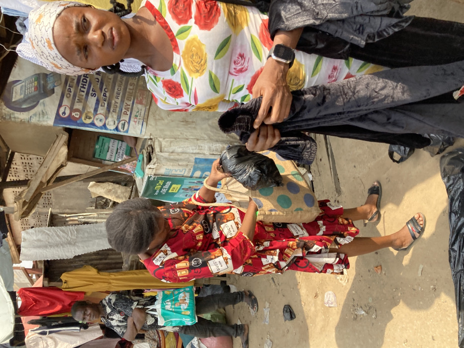 This image depicts an outdoor market scene in Ura Nta Umuarandu, Nigeria, under bright daylight.
Three individuals are visible. In the foreground right, an adult female with dark braids and a white patterned headwrap wears a white, long-sleeved shirt with a red and yellow floral pattern. She holds multiple pieces of black fabric, possibly velvet, in her hands, appearing to inspect them. A dark digital watch is present on her left wrist. Her gaze is directed towards the left side of the frame.
In the mid-ground, slightly to the left, another adult female is bent over, looking into a light brown tote bag with blue polka dots. She wears a red, long-sleeved dress featuring a repeating pattern of "GUCCI Bloom" text and what appear to be perfume bottles. A black plastic bag is held in her right hand above the tote bag. She is wearing dark flat sandals.
In the background left, a young male or teenager stands with crossed arms, looking towards the camera. He wears a short-sleeved patterned shirt and dark trousers, with dark sandals. He holds a light green tote bag with an illustrated design in his left hand.
The environment consists of unpaved, sandy ground with scattered debris, including small pieces of paper and black plastic bags. Makeshift market stalls are visible in the background, constructed from wood, corrugated metal sheets, and draped fabric. Hanging clothing items, including a red top, a yellow dress, and other garments, are displayed for sale. Prominently displayed signage on the right includes a blue and white poster depicting a POS terminal and listing services such as "OPEN ACCOUNT," "WITHDRAW," "SEND MONEY," "DEPOSIT," "BUY RECHARGE," and "PAY BILLS" (including DStv and GOtv). A smaller green and white sign below it states "ATM Card - ONE Minute." Another partial sign above includes "PAY WITH TRANSFER" and a phone number.