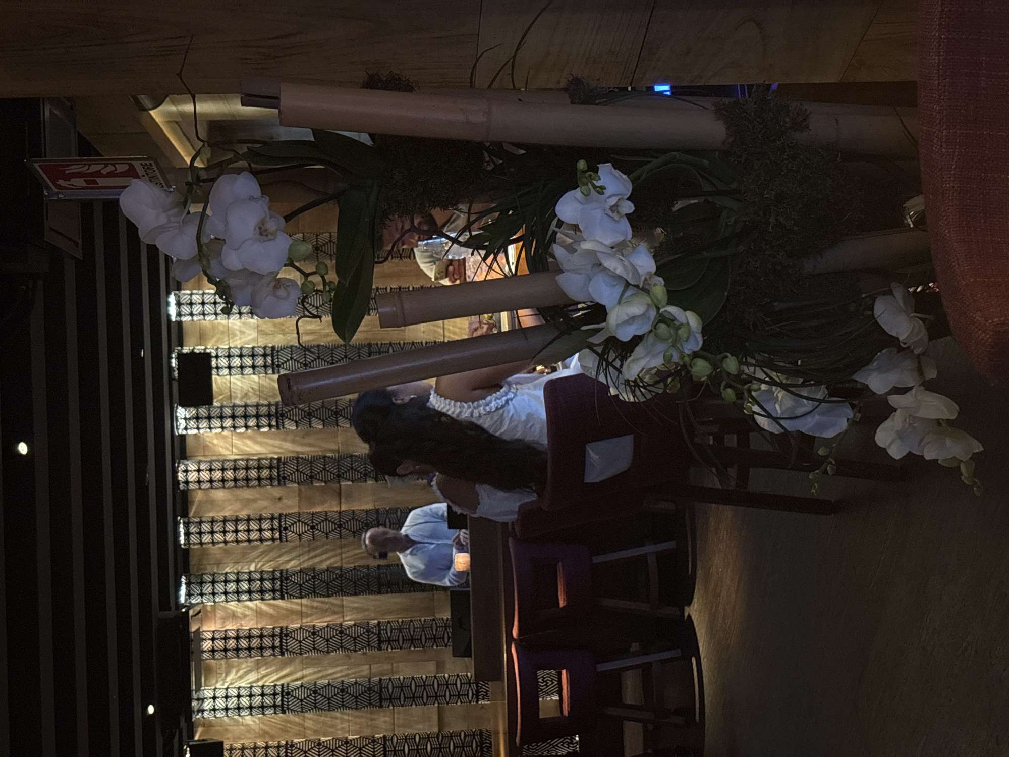 The image captures an indoor scene, possibly a restaurant or lounge, with a dimly lit ambiance. In the foreground, a large arrangement of white orchids and bamboo stalks is visible. Behind this floral display, a man in a light blue shirt stands behind a counter, seemingly attending to something. To his right, two women are seated, with one facing away from the camera and the other partially visible, also seated. The background features a wall with a textured pattern of vertical stripes, some of which are illuminated. Several dark red chairs are arranged in the seating area. A fire extinguisher sign is mounted on a wooden panel to the right of the frame. The overall impression is one of a stylish, contemporary establishment. The city of Rome, Italy, is not explicitly depicted or implied by any visual cues in the image.