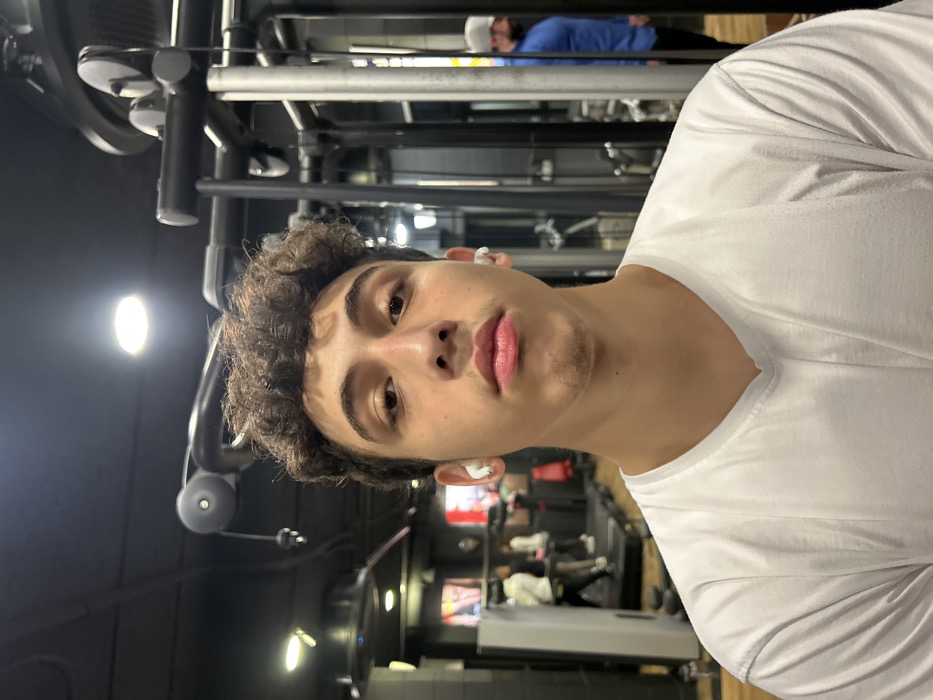 A young man with curly hair and light skin is looking at the camera. He's wearing a white t-shirt and what appear to be wireless earbuds. The image seems to be taken in a gym, with exercise equipment visible in the background. Another person, wearing a blue shirt and a white cap, is also visible, possibly working out. The lighting suggests an indoor setting with artificial lights.