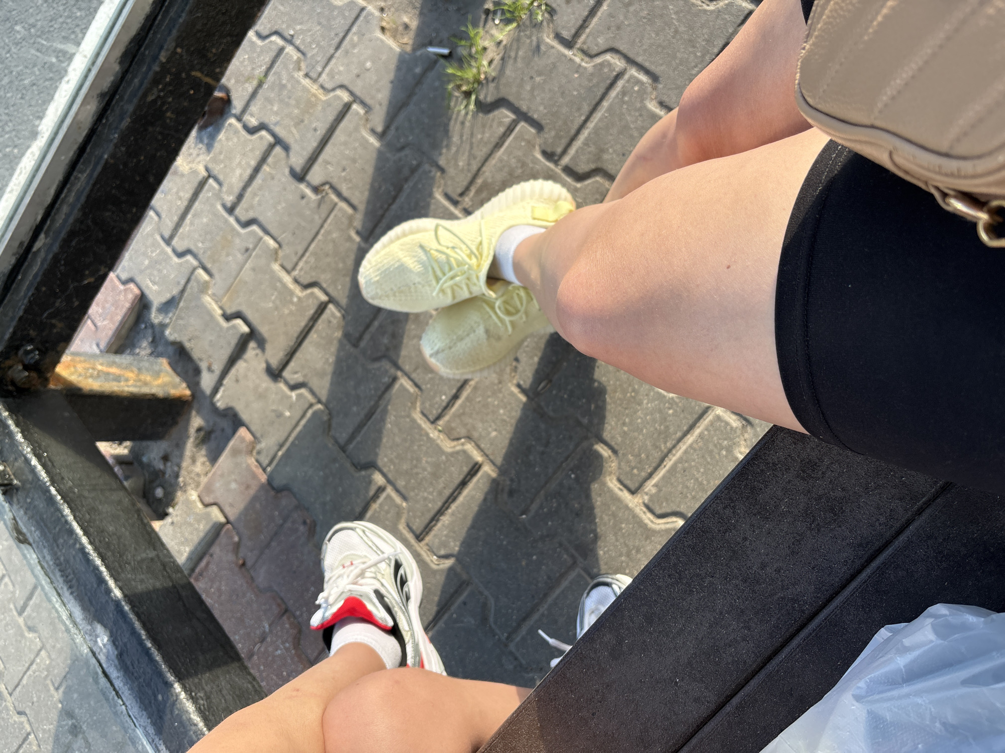 The image shows a close-up of two people's legs and feet, sitting on a textured dark surface. One person is wearing light yellow sneakers with white socks and black shorts. The other person is wearing white and red athletic shoes with white socks. The background is a grey paved surface, suggesting an outdoor setting. Sunlight casts shadows, indicating it is daytime. The image has a casual and relaxed feel. No visible text is present in the image. The location context provided is Mytishchi, Russia.