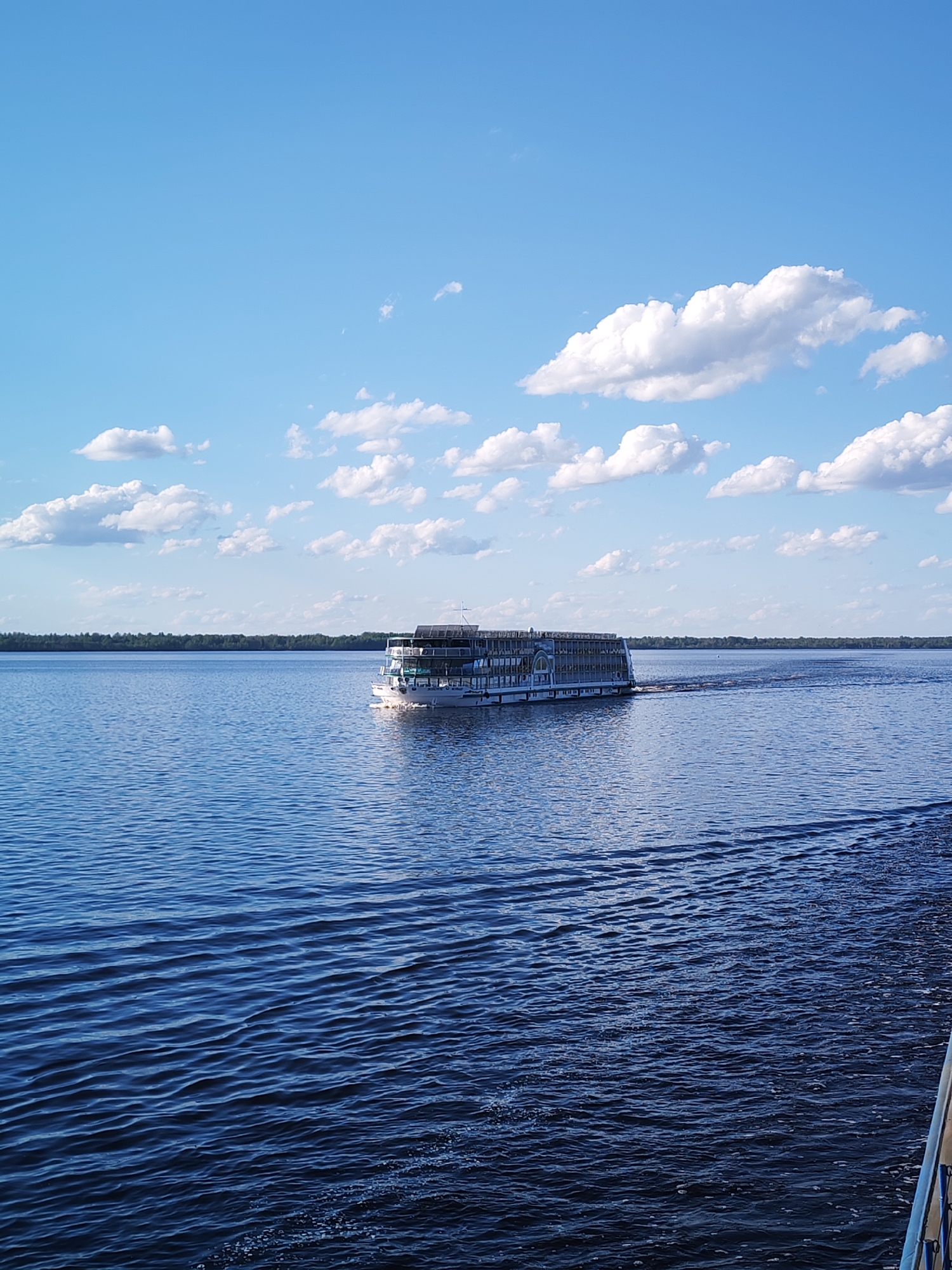 A medium shot depicts a large river passenger ship moving across the blue waters of the Volga River in Myshkin, Russia, beneath a blue sky dotted with puffy white clouds. The ship, centrally positioned, appears to be a multi-deck vessel with a white and grey exterior. It is oriented horizontally and moving to the right of the frame, leaving a wake behind it. In the background, a distant line of green trees marks the opposite shore of the river. The water surface exhibits gentle ripples and wave patterns reflecting the blue sky. The lower right corner shows a partial view of the railing of another vessel, suggesting the photo was taken from a moving boat.