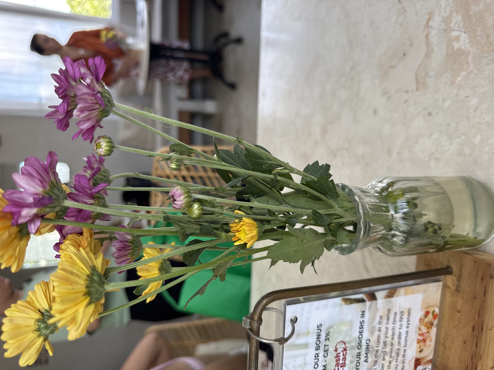 The image shows a glass vase of purple and yellow flowers sitting on a table, likely in a restaurant. A wooden stand with a sign for Amino is visible in the foreground. The sign advertises a bonus program and asks customers to fill out a form with their name. In the background, blurred people can be seen sitting at tables, including a child with an orange shirt and another table with a person sitting at it. The scene appears to be indoors, with natural light coming in from a window or opening, and is likely located in Ungasan, Indonesia due to the sign referencing the Amino app, which is common in the region.