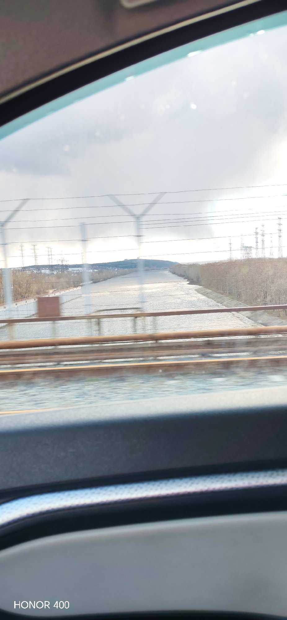 The image displays a wide-angle view of a vast body of water, likely a river or reservoir, observed from inside a moving vehicle. Elements of the vehicle's interior, including what appears to be a dashboard and a window frame, are visible at the top and bottom of the frame. In the mid-ground, the structure of a bridge is evident with horizontal railings and railroad tracks extending across the foreground.
The expansive water surface, possibly a part of the Kama River or Votkinsk Reservoir in Chaikovsky, Russia, reflects a bright, overcast sky, giving it a shimmering appearance. Both banks of the water are lined with trees, mostly bare, suggesting the season is late autumn, winter, or early spring. Numerous tall electricity pylons (power line towers) are scattered across the landscape on both sides of the water, indicating extensive power infrastructure. A distant landmass or low hill is visible on the left horizon. The sky is mostly cloudy, contributing to a somewhat diffused lighting, though there are bright areas suggesting the sun is partially breaking through or hidden behind light clouds. The overall scene depicts a blend of natural and industrial elements. The text "HONOR 400" is visible in the bottom left corner of the image.