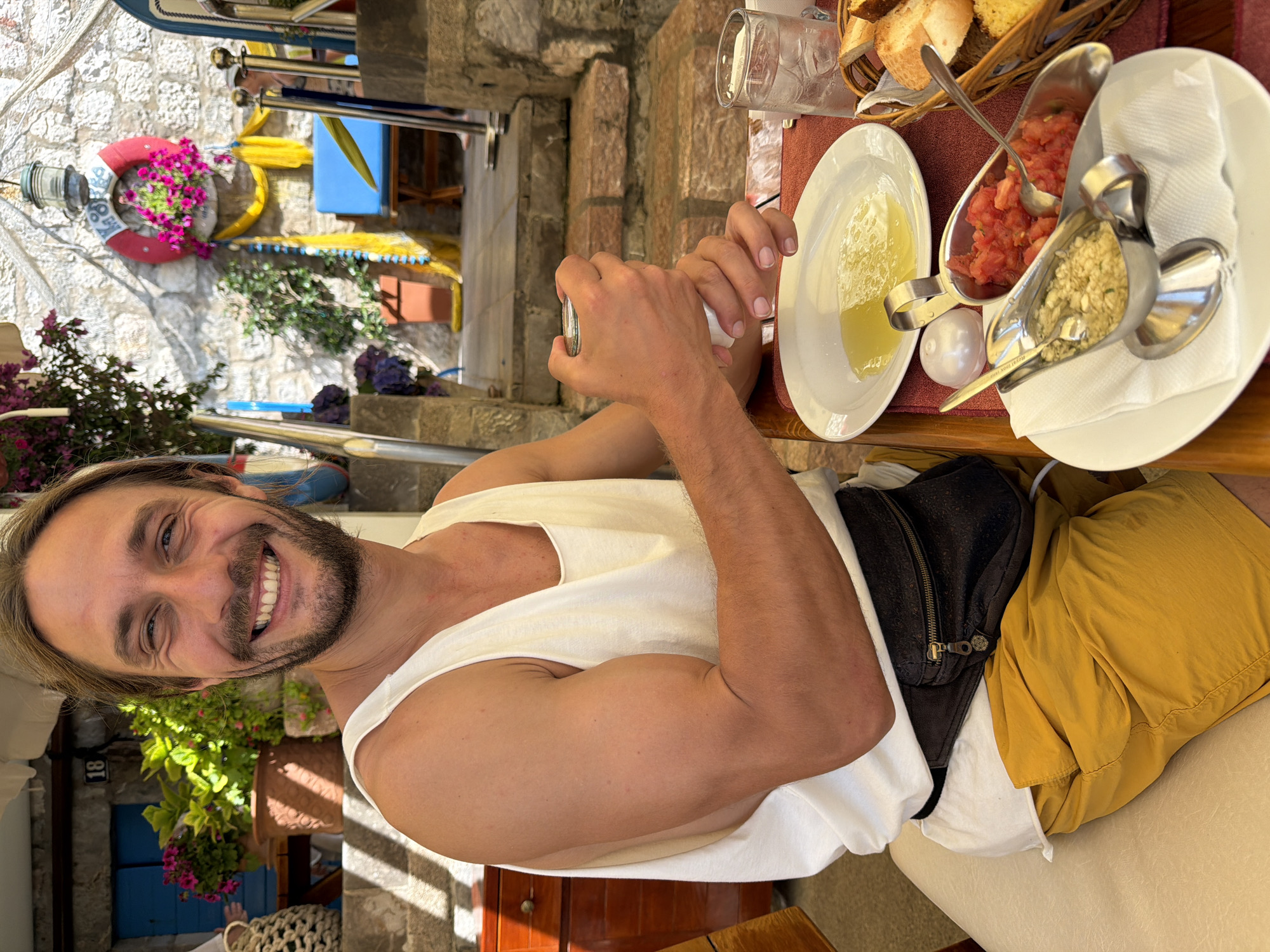 A smiling man with blonde hair and a beard sits at a table with food and drinks. He is wearing a white tank top and yellow shorts, with a fanny pack around his waist. In front of him is a white plate with a dish of food, a glass of water with ice, a basket of bread, and two small metal dishes containing chopped tomatoes and a finely chopped mixture of garlic and herbs. The man is holding a small metal container in his hands.
The setting appears to be an outdoor restaurant or cafe, with stone walls and steps in the background. There are potted plants and flowers visible, adding to a pleasant ambiance. A red and white life preserver with flowers hangs on the wall, along with a yellow cloth draped behind it. The overall scene suggests a relaxed meal in a sunny location. This scene is likely in the city of Pržno, Montenegro, based on the architectural style and lush vegetation typical of the area.