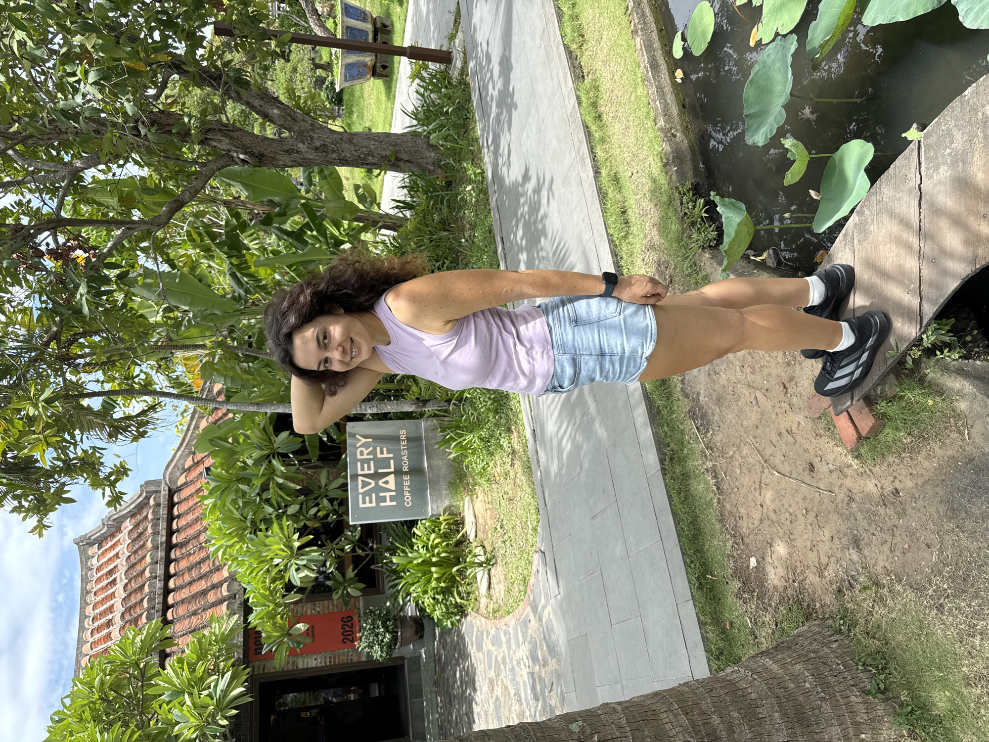 A young woman stands in front of a cafe named "EVERY HALF COFFEE ROASTERS" in Hoi An, Vietnam. She is wearing a light purple tank top and denim shorts, with black athletic shoes. Her left arm is raised behind her head, and she is smiling at the camera. The cafe has a traditional Vietnamese tiled roof and a brick facade. In the background, there are lush green trees and a pond with lily pads. The ground is paved with gray stone tiles and dirt. The woman is standing on a wooden plank that extends over the pond. The cafe appears to be situated in a garden-like setting.