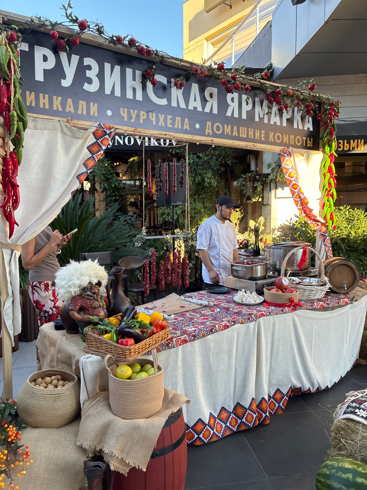 The image depicts an outdoor food stall in Sochi, Russia, specializing in Georgian cuisine. The stall features a prominent sign with Russian text, "ГРУЗИНСКАЯ ЯРМАРКА" (Georgian Fair), and also lists "ХИНКАЛИ" (khinkali), "ЧУРЧХЕЛА" (churchkhela), and "ДОМАШНИЕ КОМПОТЫ" (homemade compotes). The stall is decorated with strings of red chilies and pomegranates, adding to the festive atmosphere. A chef in a white uniform stands behind a table laden with food items, including pots on a portable stove, baskets of fruits and vegetables like eggplants, peppers, and lemons, and strings of dried meat or churchkhela hanging in the background. A woman in casual attire is visible to the left, looking at her phone. The stall has a traditional Georgian aesthetic, with patterned textiles and rustic elements like barrels and woven baskets. The overall scene suggests a lively market setting, likely during daytime, with a focus on authentic Georgian culinary offerings.