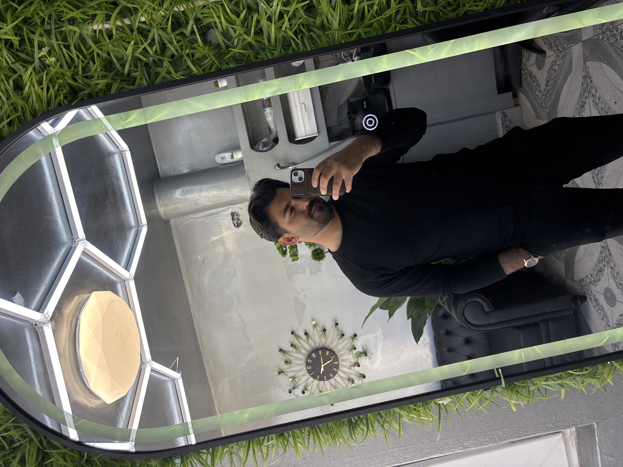 A man takes a selfie in a mirror. The mirror is surrounded by artificial green grass. The environment reflected in the mirror includes a gray wall, a black chair, and a decorative wall clock. The man is wearing a black sweater and holding a phone. The location of the photo in Dolni Tsibar, Bulgaria is not discernible.