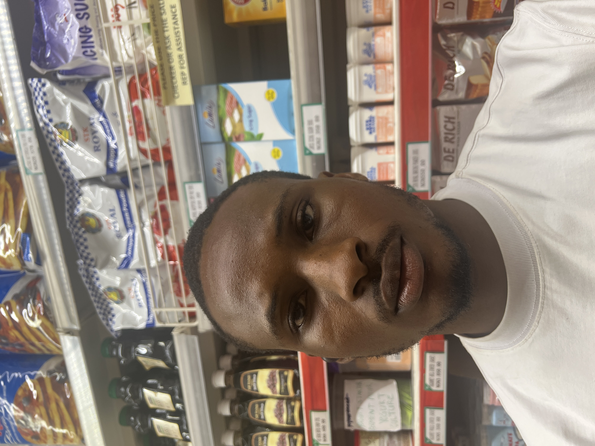 A man is in a grocery store, standing in front of shelves stocked with various food items. The shelves contain packages of Royal brand whipped cream powder, sugar, bottles of olive oil, yogurt culture, and De Rich high sugar. Price tags are visible on the shelves. The man is wearing a white t-shirt. The location is in Abuja, Nigeria.