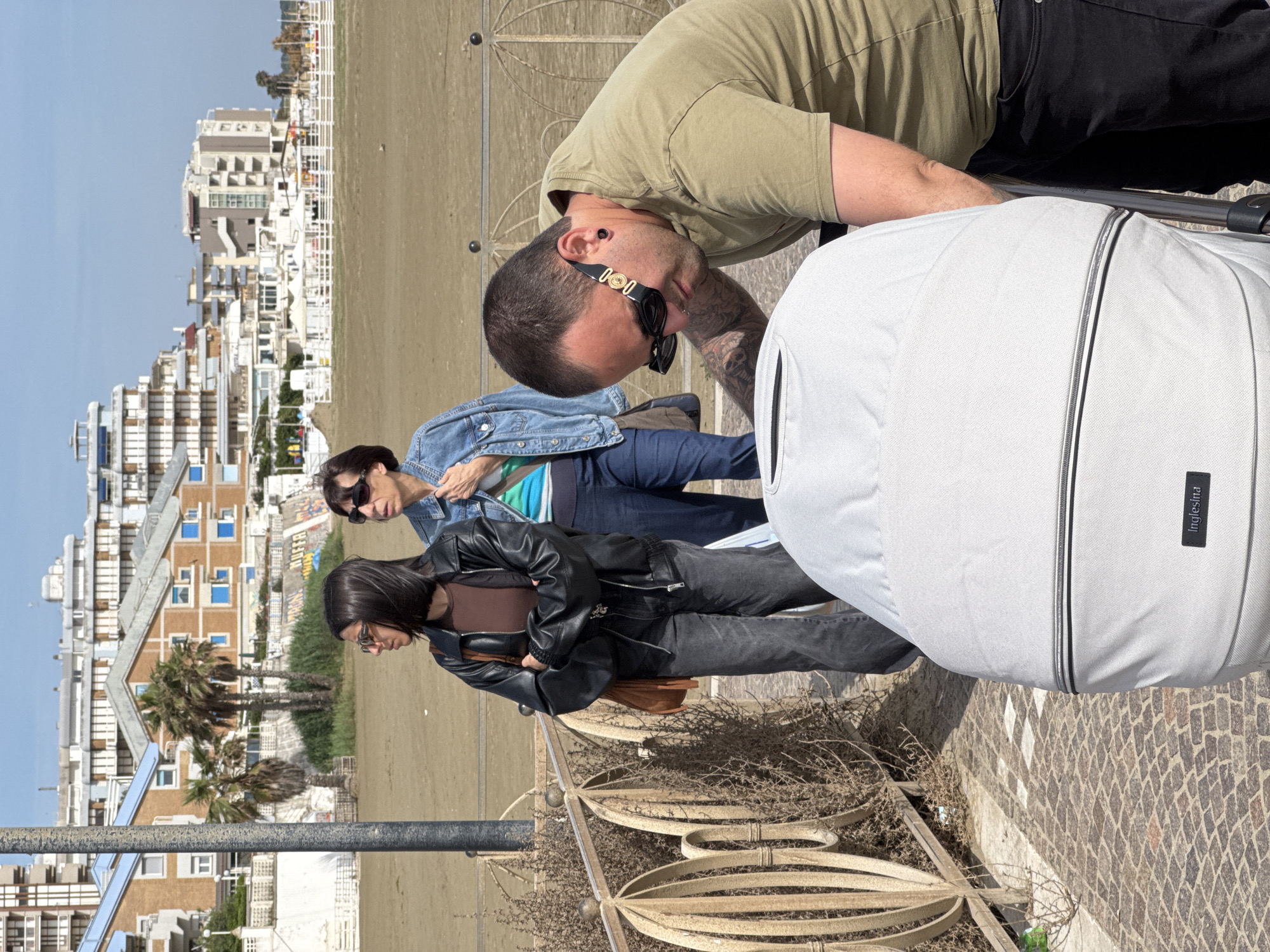 The image shows a group of people on a paved promenade overlooking a beach. In the foreground, a man wearing sunglasses, a light green shirt, and black pants is leaning over a white stroller. Two women are standing behind the man, looking in the same direction. One woman is wearing a denim jacket and jeans, while the other is wearing a black leather jacket, brown top, and dark jeans.
The background features a beach with white sand and a row of buildings in the distance. Palm trees are visible near the buildings.
The location is Nettuno, Italy.