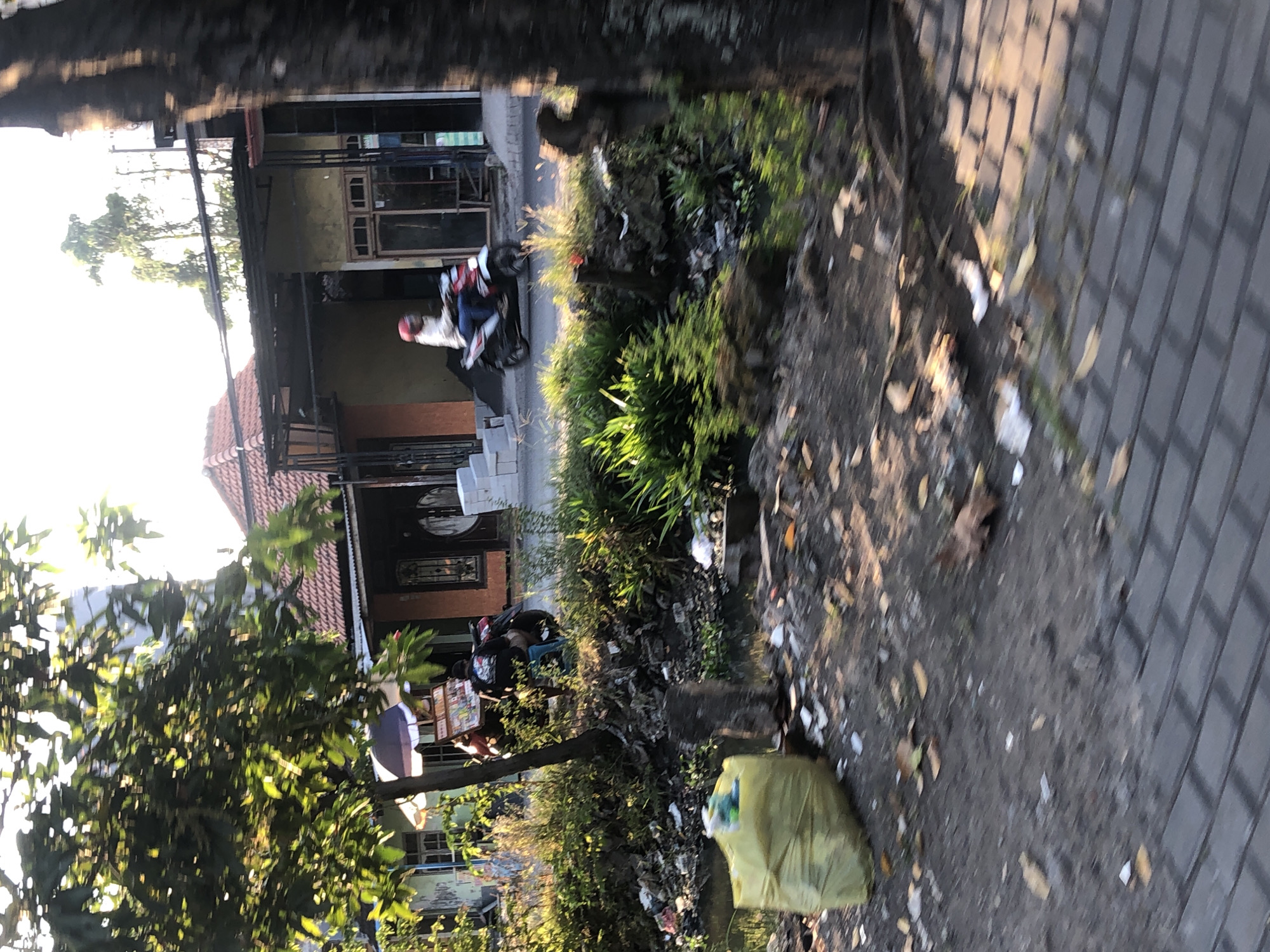 The image depicts a street scene in Kalidawir, Indonesia, characterized by a mix of residential buildings, vegetation, and visible trash. In the foreground, a bright yellow trash bag is prominently placed on the ground next to a waterway. Further into the scene, a person is riding a motorcycle, heading towards the right of the frame. Behind the motorcycle, a small food cart with an umbrella is visible, suggesting a commercial area. The buildings appear to be single-story structures with tiled roofs. The overall environment shows signs of neglect, with debris and vegetation accumulating around the waterway. The lighting suggests it is late afternoon or early morning, with the sun casting long shadows.