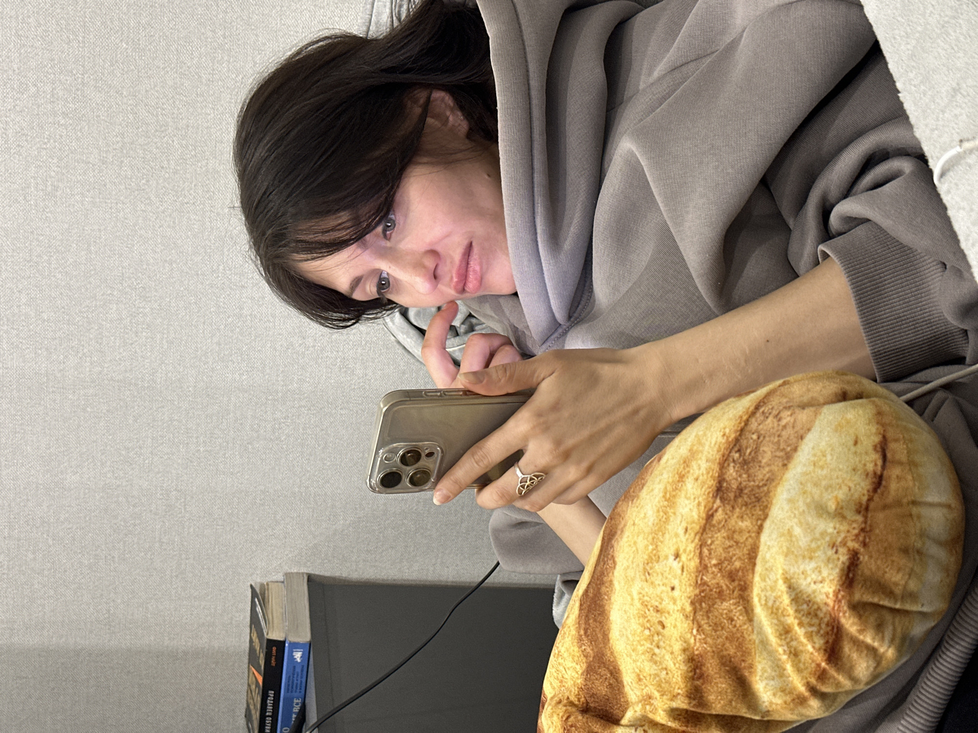 A young woman, likely in her 20s or 30s, is pictured in a relaxed, intimate indoor setting in Murino, Russia. She is reclined, possibly on a sofa or bed, and appears engrossed with her gold-colored smartphone, which she holds in her right hand. Her dark hair frames her face, and her gaze is directed towards the phone screen, while her left hand gently touches her chin or cheek. She wears a comfortable, oversized grey hooded sweatshirt. A silver-colored ring with an intricate, knot-like design is visible on her right ring finger.
A distinctive pillow or blanket, printed with a realistic image resembling a loaf of toasted bread or brioche, is partially visible on the right side of the frame. Below the smartphone, a small stack of books includes one with a blue spine showing the partial text "RICE" and another with a yellow/orange spine where the Russian word "ПРОДАВЕЦ" (salesman) is partially legible. A black cable is also visible near the books. The background consists of a plain, light grey textured wall, suggesting a simple and cozy environment. The soft, ambient lighting points to an indoor scene, likely during the evening. Her expression appears calm and focused.