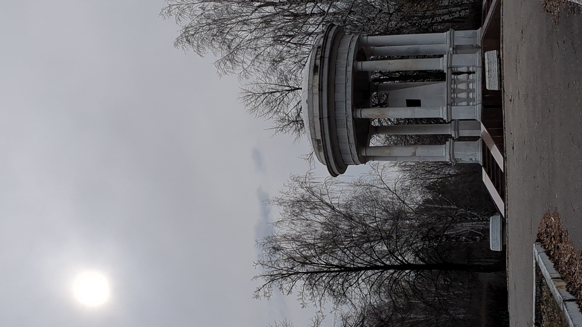 The image shows a rotunda in Ulyanovsk, Russia. The structure is made of gray stone and consists of a circular base, columns, and a dome-shaped roof. Bare trees surround the rotunda. To the right, there is a paved path or road and a low wall. The sky is overcast and the sun is visible low in the sky.