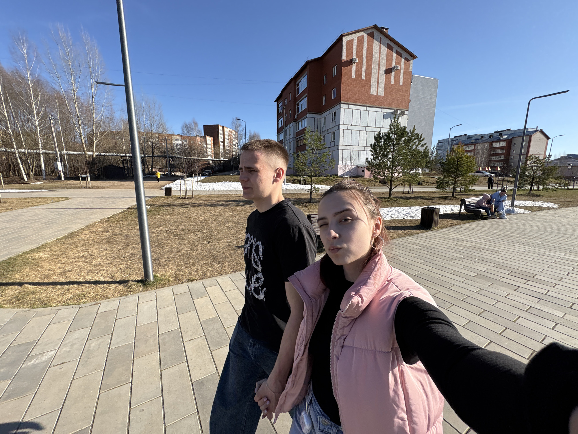 The image depicts a bright, sunny outdoor scene, likely a park or residential area in Votkinsk, Russia. Two young adults are prominently featured in the foreground, taking a selfie while walking along a paved pathway.
On the left, a young man with short, light brown hair wears a black t-shirt with a white graphic design and dark jeans. He looks towards the left, with a neutral expression. To his right, a young woman, holding the camera for the selfie, wears a pink puffer vest over a black long-sleeved shirt and jeans. She gazes directly at the camera with a playful, slightly pouting expression. Their hands are clasped together, suggesting a romantic relationship.
The background reveals a clear blue sky and signs of early spring, with patches of melting snow on the ground. Bare deciduous trees, possibly birches, line the left side, while evergreen trees are scattered closer to the buildings. Several multi-story residential buildings are visible; one prominent building on the right features red brick with white vertical architectural accents and air conditioning units. Other grey and brown apartment blocks are seen in the distance. Horizontal pipes run across the mid-ground on the left.
Further into the scene, two people are seated on a park bench, one in light blue clothing and the other in a darker top. Another individual in a yellow jacket can be seen walking in the distance. The strong shadows indicate bright daylight. No discernible text is visible in the image. The scene conveys a casual, pleasant outing on a sunny day.