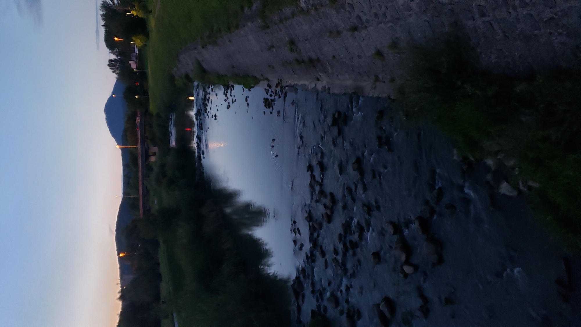 The image depicts a river flowing through a park or natural area at dusk. The sky is a gradient of deep blue to a lighter hue near the horizon, suggesting twilight. A prominent mountain is visible in the distance, with lights on its summit, possibly indicating a communication tower or a distant settlement. Several streetlights are visible along the riverbanks and near a bridge in the midground. The bridge appears to be a road bridge, with traffic lights or vehicle lights visible on it. The river has a rocky bottom, with water flowing over the stones. The banks are lined with grass and trees, some of which are silhouetted against the sky. The scene is peaceful and natural, with evidence of urban infrastructure in the background. The specific location in Prešov, Slovakia, is identifiable by the distinctive mountain and the river's characteristics, potentially the Torysa River, which flows through the city.