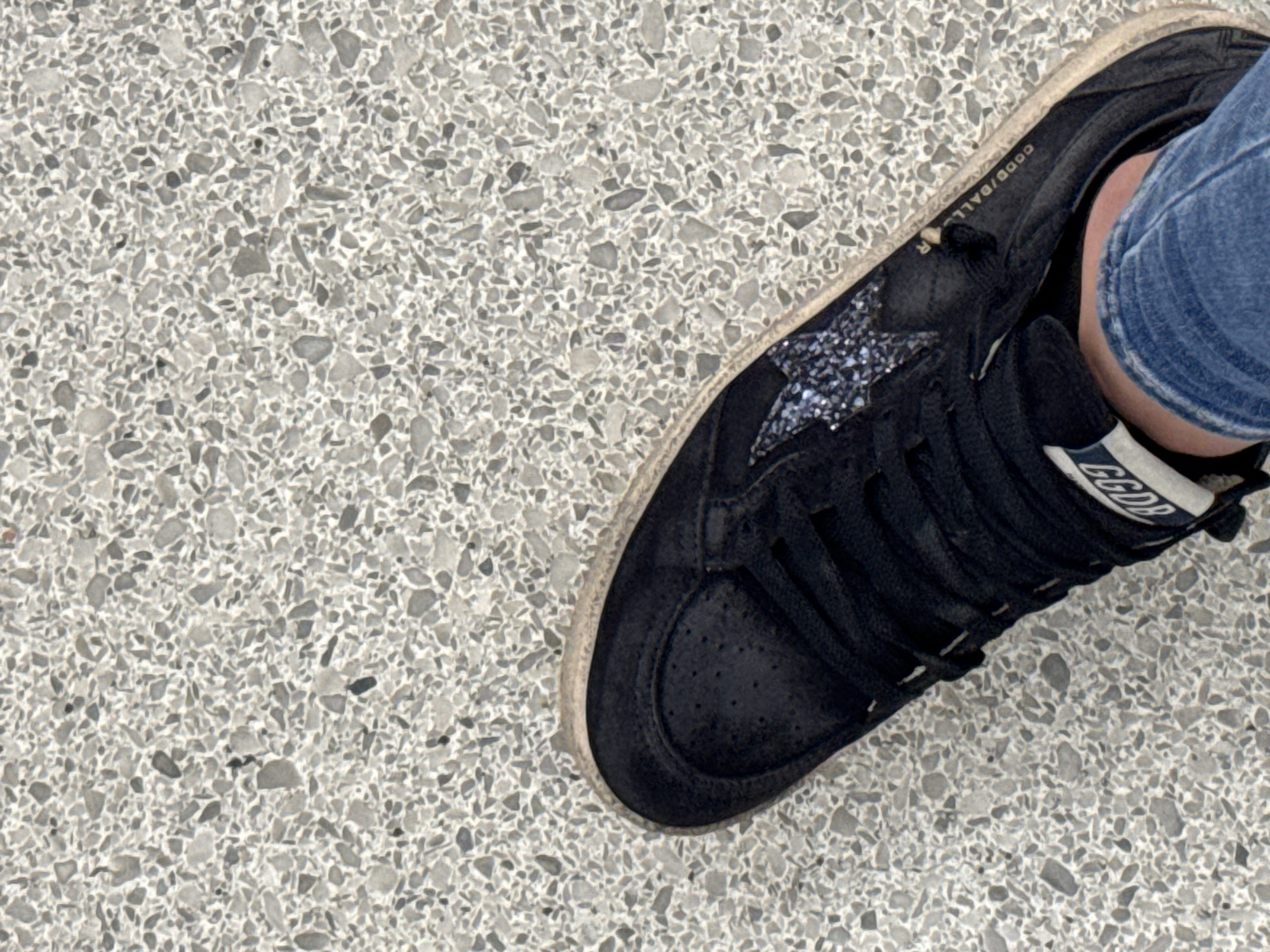 The image depicts a close-up view of a person's foot wearing a black Golden Goose sneaker with a glittery star detail, resting on a terrazzo floor. The person is wearing blue jeans. The floor appears to be a light gray color with small aggregate pieces. The location is likely within San Antonio, Texas, USA, based on the user's request.