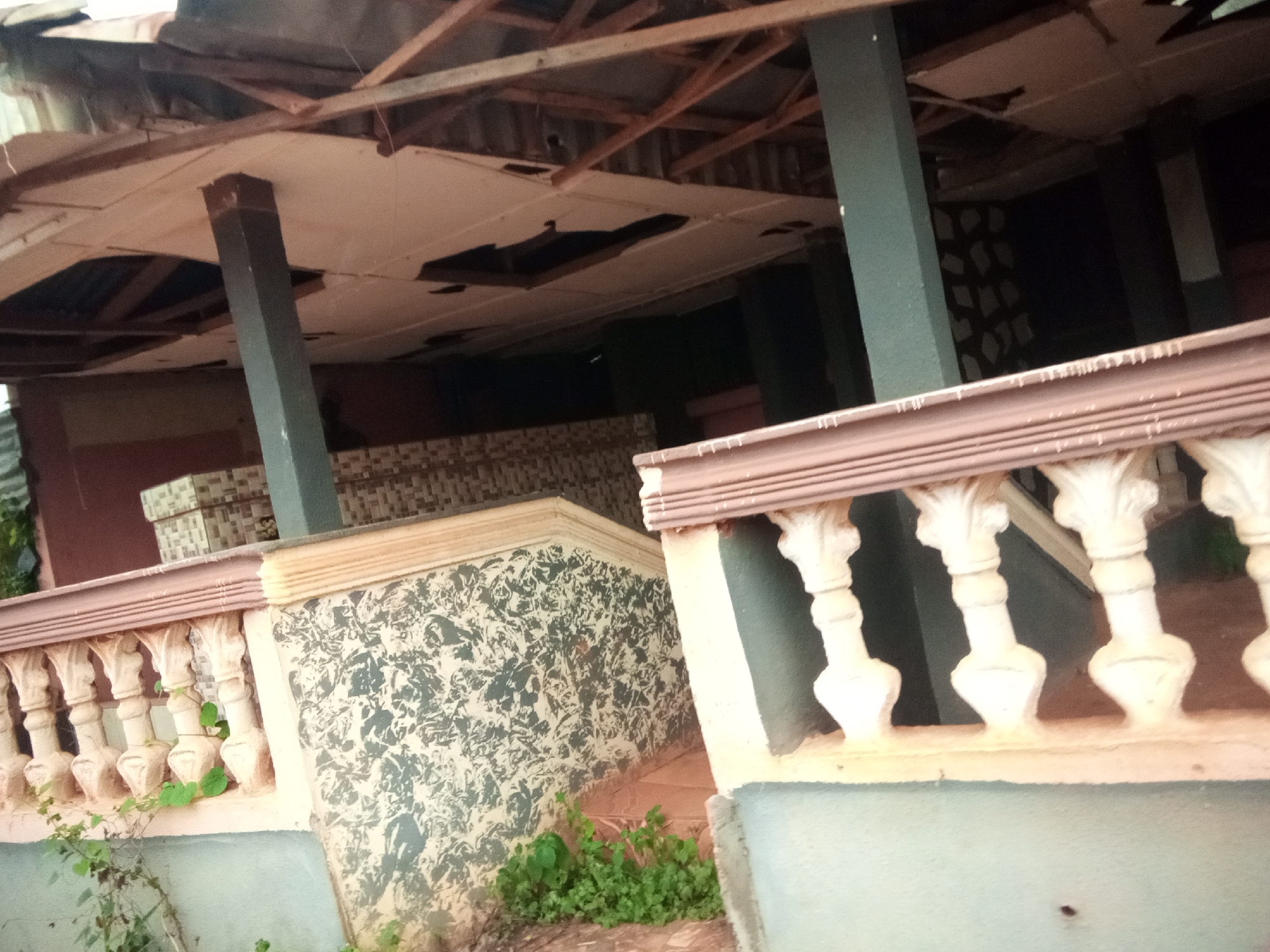 The image depicts a partially collapsed outdoor structure in Oko Town, Nigeria. The roof shows signs of disrepair with exposed wooden beams and missing ceiling panels. Pillars, some of which appear to be made of concrete, support the damaged roof. A decorative railing with white balusters runs along a raised platform, with climbing plants growing at its base. The wall of the platform is decorated with a patterned design. In the background, stacks of what appear to be boxes or building materials are visible. The overall scene suggests an abandoned or neglected building.