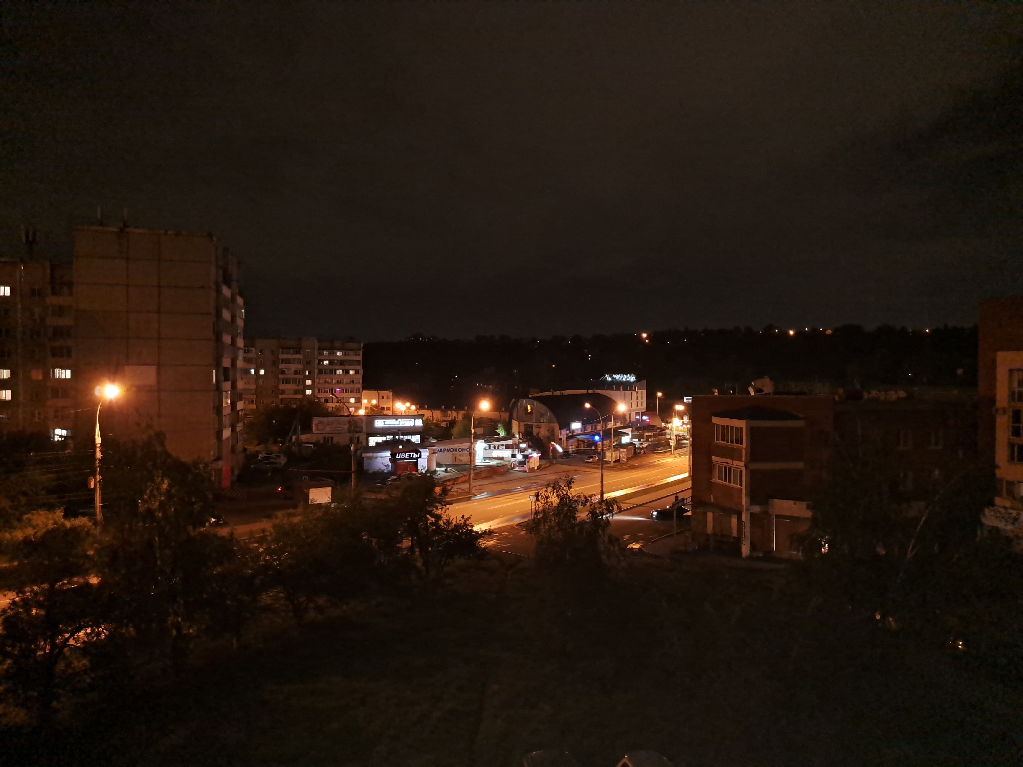 This is an aerial, night-time, long-exposure photograph of a city intersection in Irkutsk, Russia. The image captures a street scene illuminated by streetlights and the lights of various buildings and vehicles. The foreground is dominated by dark foliage, likely trees, obscuring the ground level.
In the middle ground, a road curves through the scene, with visible lane markings and streetlights casting pools of light. Several buildings line the road. On the left, a multi-story apartment building is visible, with illuminated windows. Adjacent to it are smaller commercial buildings with illuminated signage, including one that reads "ЦВЕТЫ" (Flowers) in Russian. Further down the road, a large, arched building with external lighting is present. To the right of the road, another multi-story brick building is visible, with a prominent section featuring multiple windows.
Vehicles are present on the road, with headlights and taillights visible, indicating movement. One car's headlights are clearly illuminated as it travels along the road. The sky is dark, typical of nighttime, with some subtle cloud cover visible. The overall impression is of an active, urban area at night, with the lights providing a stark contrast to the darkness.