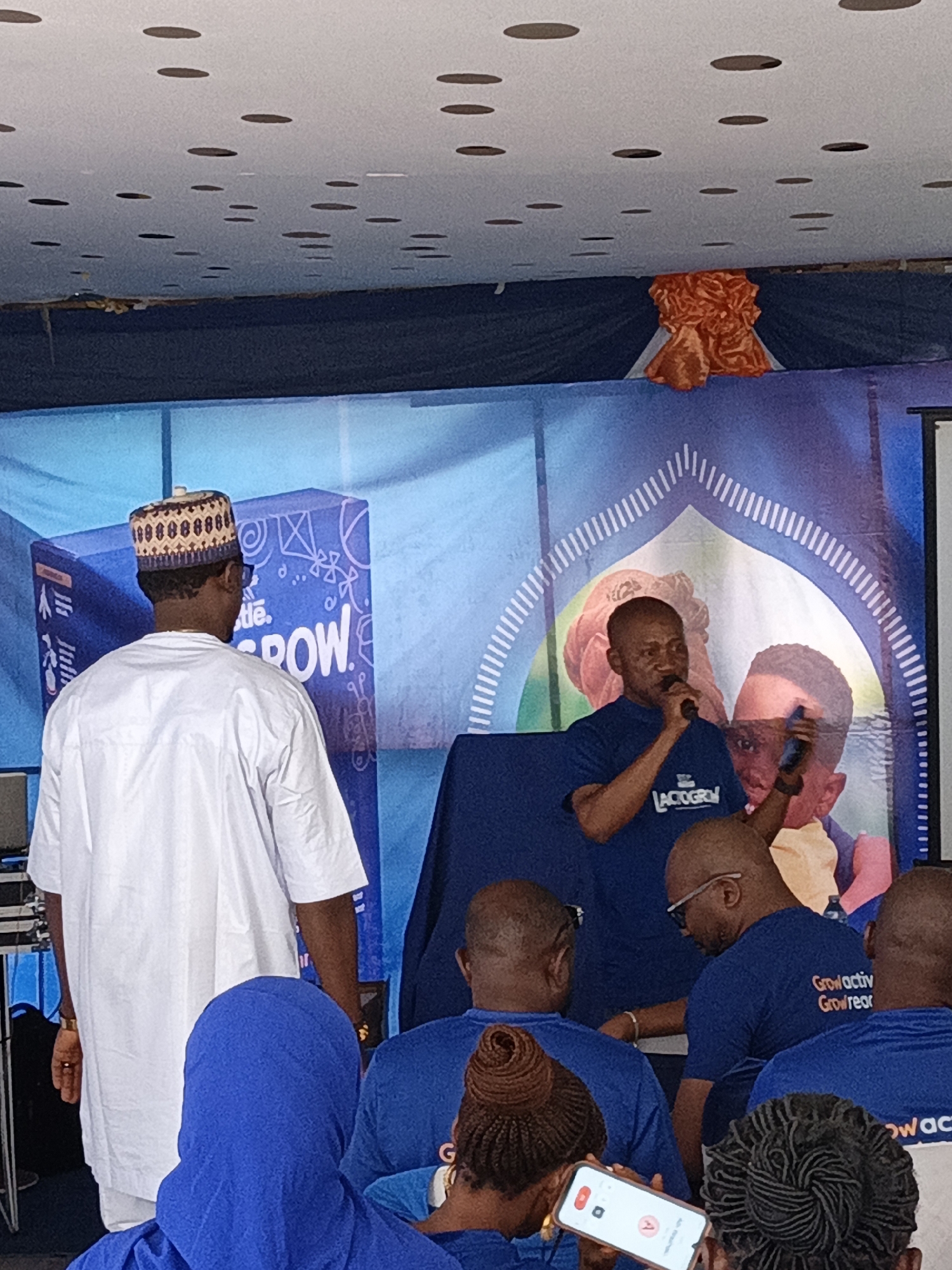 The image captures an event likely held in Tarauni, Nigeria, focused on a product called "LACTOGROW." A man in a blue shirt stands at a podium speaking into a microphone, with a "LACTOGROW" logo visible on his shirt. Behind him is a backdrop with the LACTOGROW brand. Seated in front of him is an audience of people wearing blue shirts with "Grow activ Grow rear" printed on the back. One person is seen holding a cell phone recording the event. A man in a traditional white outfit and cap is standing to the side. The environment suggests an indoor venue with decorations.