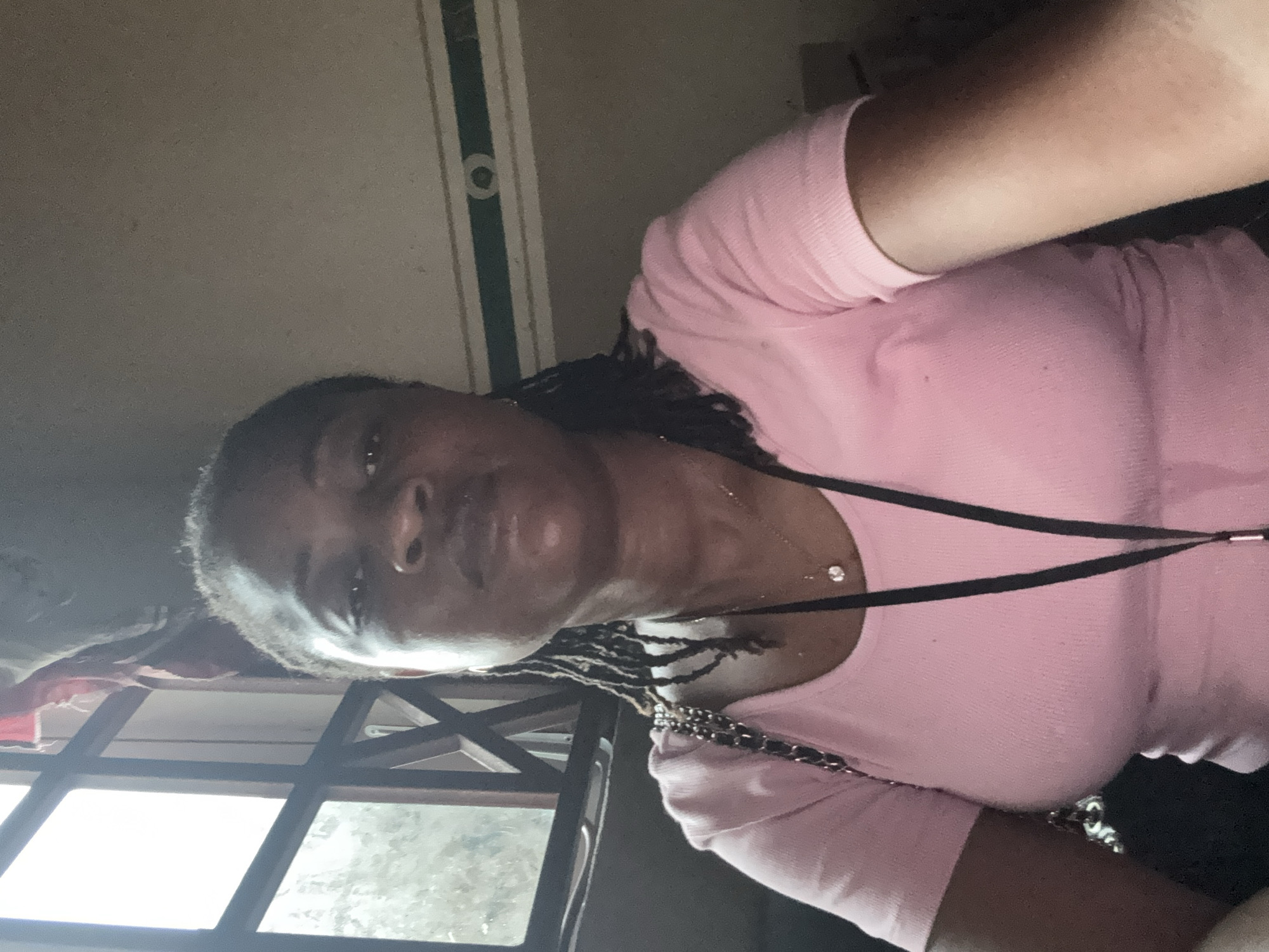 A woman, facing the camera, with dark skin and braided hair is wearing a pink t-shirt. She is wearing a necklace with a diamond pendant. The background appears to be indoors, with a ceiling and a window with a view of foliage and what appears to be a grid-like structure. The lighting suggests it might be daytime. The overall tone of the image is casual.