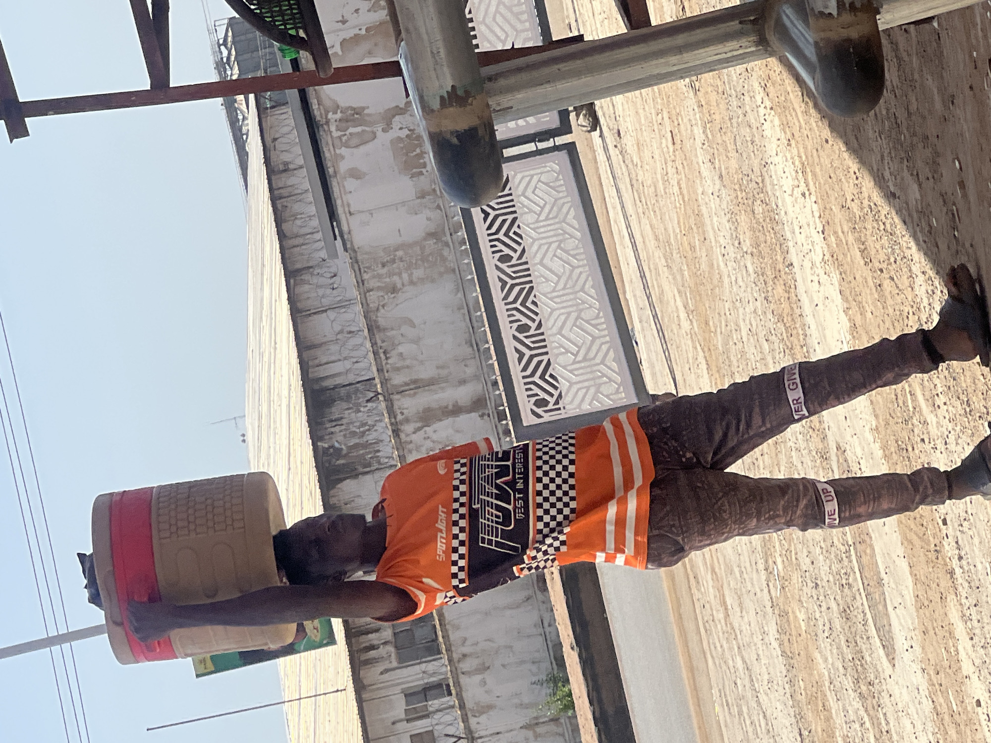 A man carries a water container on his head, in an outdoor urban environment. The man is wearing an orange t-shirt with black, white, and gray graphics, and long brown pants with the phrase "Never Give Up" repeated in white letters on a black strip around each leg. He is wearing black shoes. He is walking along a sidewalk or street in front of tall, nondescript buildings. Overhead are metal pipes and a metal lattice structure. The buildings appear aged and worn. The setting is suggestive of Barakin Lerawi, Nigeria.