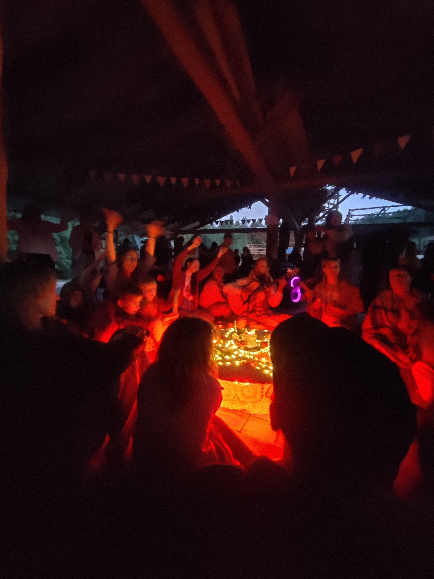 A group of people are gathered under a covered structure at night. The scene is illuminated by red and orange lights, casting a warm glow. In the center of the gathering, there appears to be a table or platform adorned with numerous string lights, possibly a decorative arrangement or a makeshift stage. Some individuals are standing with their arms raised, suggesting dancing or active participation. Others are seated, facing the center. A colorful, illuminated object, possibly a speaker or a decorative light in the shape of a number "8", is visible among the seated individuals. The background is dark, hinting at an outdoor setting, possibly a rural area or festival. The location is identified as Izky, Ukraine.