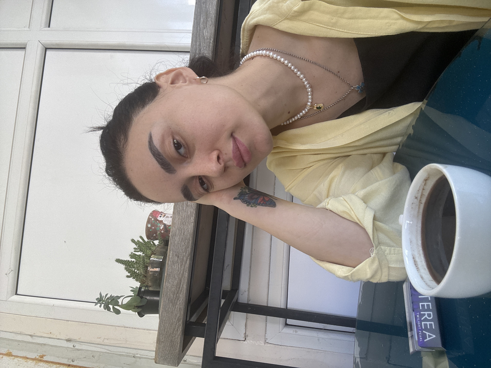 A young woman poses at a cafe table, likely outdoors, in Tbilisi, Georgia. She is resting her head on her left hand and looking at the camera. A tattoo is visible on her left forearm. She wears a yellow jacket over a black top and a pearl necklace. A white mug, presumably containing coffee, sits on the table next to a package of "TEREA" Twilight Pearl cigarettes. The table has a dark blue surface. A window is visible behind the woman, and a small collection of potted plants sits on a shelf in front of it.