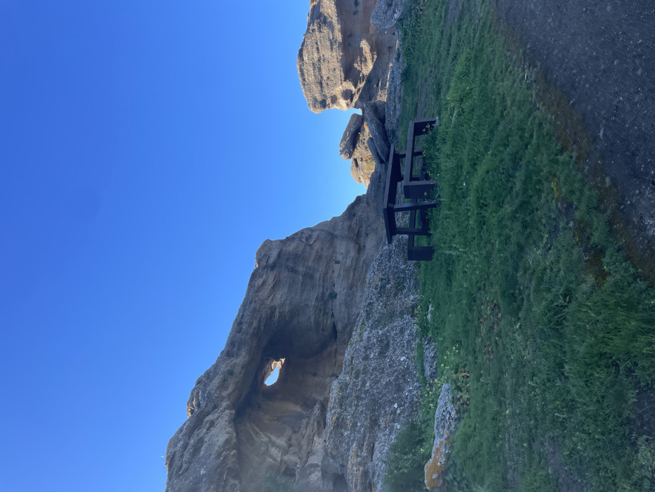 The image shows a wooden picnic table situated on the grassy slope of a rocky outcrop in Álora, Spain. The coordinates provided place the location at the base of the rock formations near the edge of a cliff with a clear view of the surrounding landscape. The table is situated under the shade of a large rock formation, with a natural archway visible through the rock. The sky is a bright blue. The environment is dry and rocky, with sparse vegetation growing on the slopes. The table appears to be empty and there are no other people or objects visible in the scene.