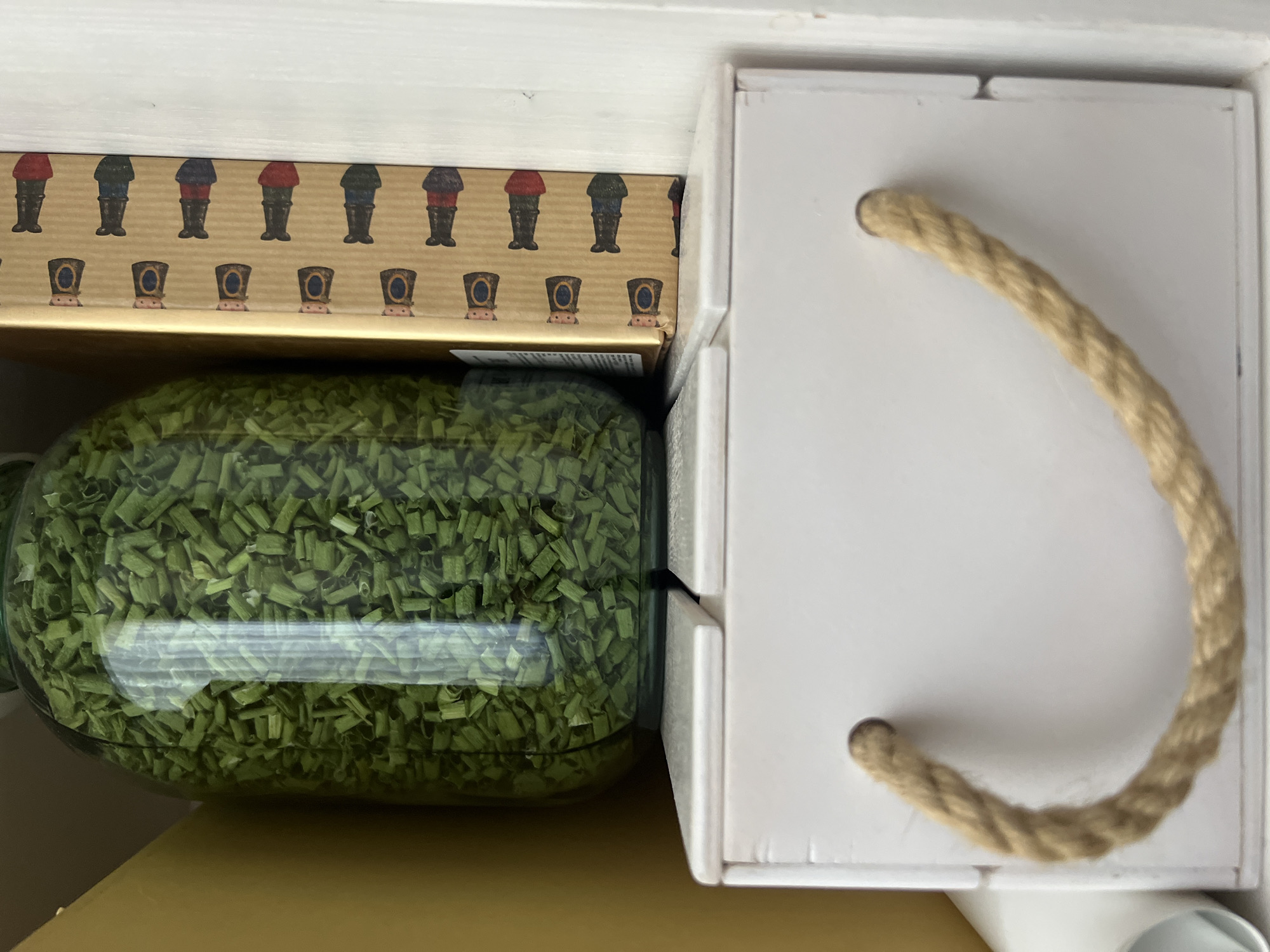 The image shows a close-up view of a shelf containing several items. On the left is a large glass jar filled with chopped green herbs, likely chives, which appears to be preserved in some liquid. To the right of the jar is a tall, rectangular cardboard box with a repeating pattern of small, colorful figures resembling chess pieces or nutcrackers. The box is golden brown in color. Below these items sits a white, rectangular wooden box with rope handles. The background consists of a yellow surface on the left and a white vertical surface on the right, suggesting a wall or cupboard. The overall setting appears to be indoors, possibly a kitchen or pantry. There are no people or actions depicted in the image. The location in Tolyatti, Russia, is not discernible from the visual information provided.