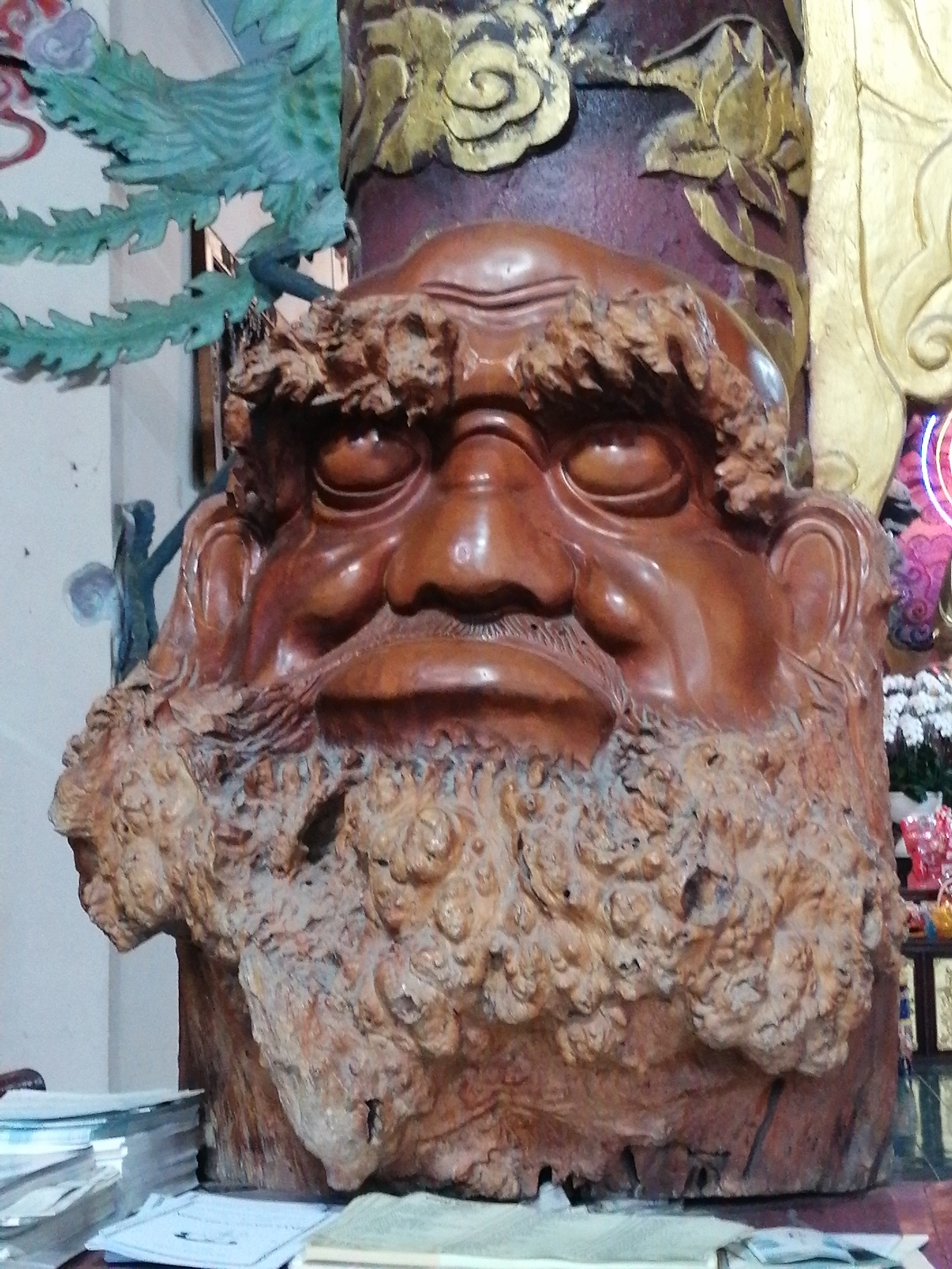 The image shows a close-up of a large, intricately carved wooden sculpture of a face with a bushy beard, situated in a room that appears to be a shrine or temple. The sculpture is predominantly made of reddish-brown wood, with textured details on the beard and hair. Behind the sculpture, parts of a larger decorative structure are visible, including gold leaf embellishments and a green, wing-like carving. In the foreground, a stack of books and papers rests on a surface. The overall impression is of a culturally significant artifact displayed in a traditional setting. Without further information or context, the specific location within Lâm Viên - Đà Lạt, Vietnam, cannot be determined.