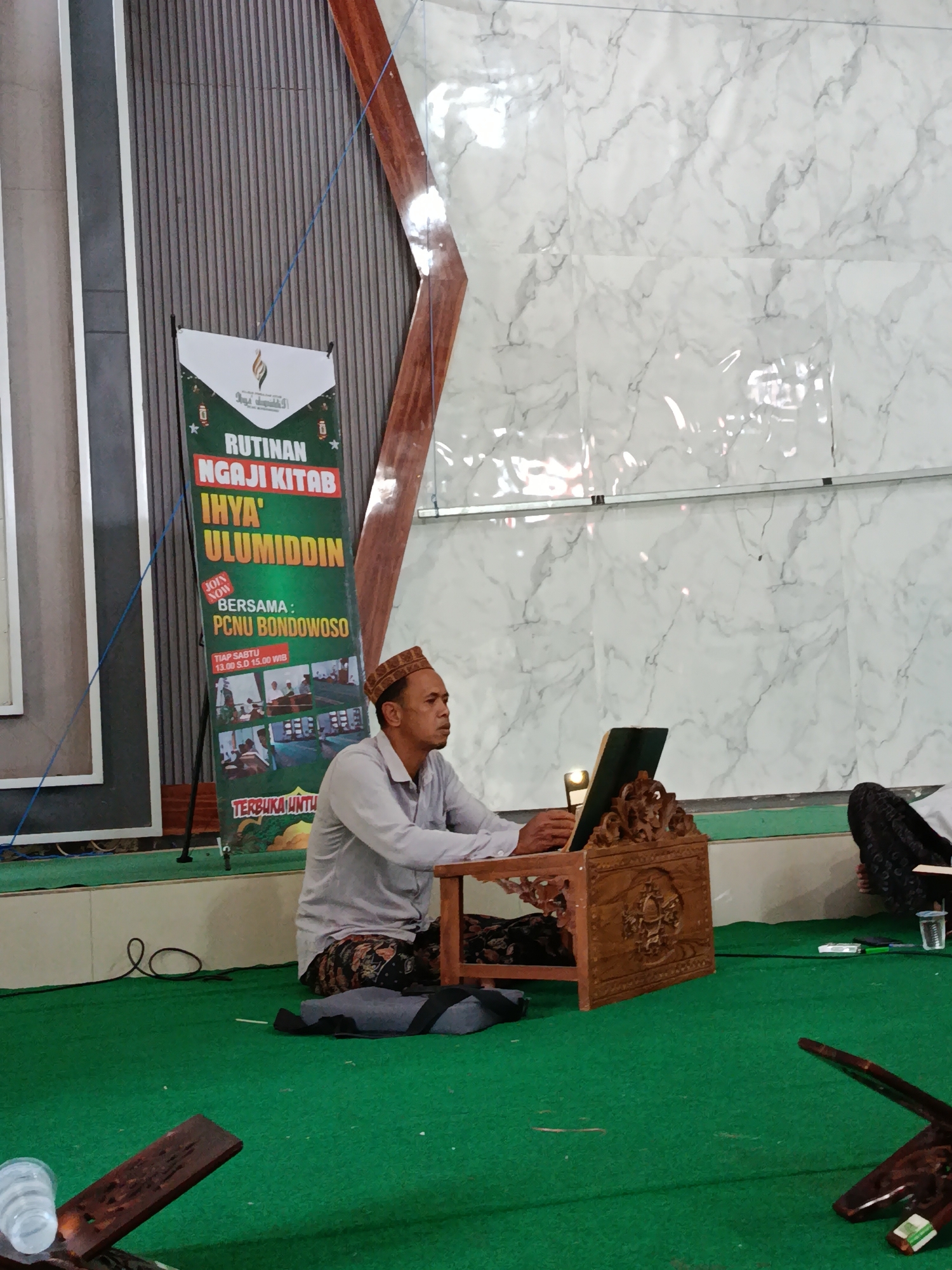 The image depicts a scene inside a building, likely a religious or community hall, in Bondowoso, Indonesia. In the foreground, a man wearing a peci (a traditional cap) and batik sarong sits on a green carpet, facing to his right. He is dressed in a light-colored shirt and appears to be engaged in reading or reciting from a book placed on a decorative wooden lectern. The lectern features intricate carvings.
Behind the man, a large banner is visible. It is green and white, with text that appears to be in Indonesian, mentioning "RUTINAN NGAJI KITAB" (regular Quranic study) and "IHYA' ULUMIDDIN" (a prominent Islamic book by Al-Ghazali). The banner also indicates "BERSAMA: PCNU BONDOWOSO" (together with: PCNU Bondowoso), suggesting the event is organized by this organization. It also lists the schedule as "TIAP SABTU 13.00 S.D 15.00 WIB" (every Saturday 1:00 PM to 3:00 PM Western Indonesian Time). The bottom of the banner has the word "TERBUKA UNTU" which likely means "Open for".
The background of the scene features a wall with a marble-like pattern. To the left of the banner, there is a wooden structure. The overall environment suggests an indoor gathering or event. In the far right, a portion of another person's body, possibly their legs and attire, is visible. The lighting appears to be artificial, illuminating the space.