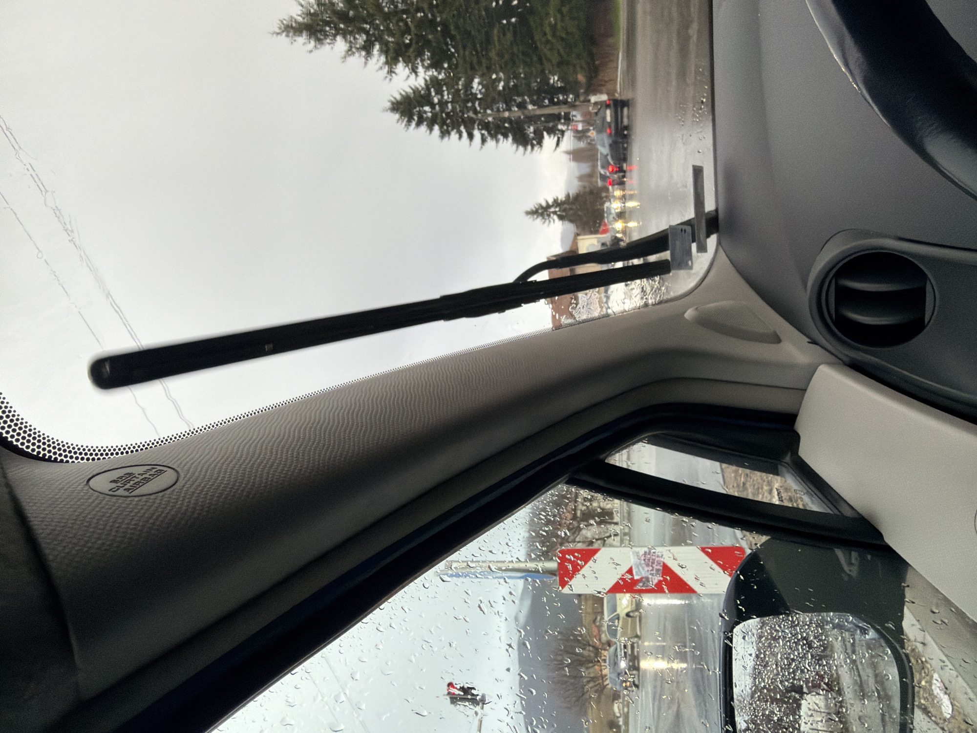 The image, taken from inside a vehicle, shows a rainy scene. The view through the windshield is partially obscured by raindrops and windshield wipers. Outside, trees are visible along with other vehicles and buildings. The side mirror is also covered in raindrops. Through the side window, a sign with red and white stripes can be seen. Inside the car, the dashboard, steering wheel, and an air vent are visible. The image was likely taken in Dragichevo, Bulgaria.